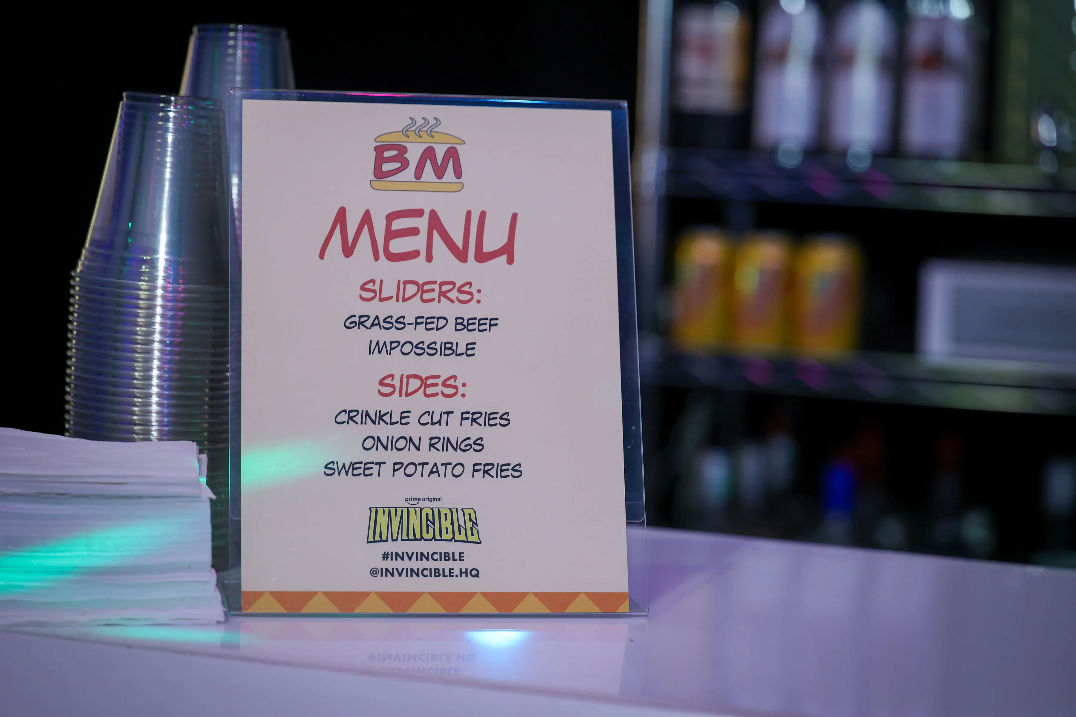 Tabletop plastic-framed menu listing sliders and sides with an Invincible logo and social tags at the bottom, stacked clear plastic cups and napkins to the left, blurred drinks cooler behind.