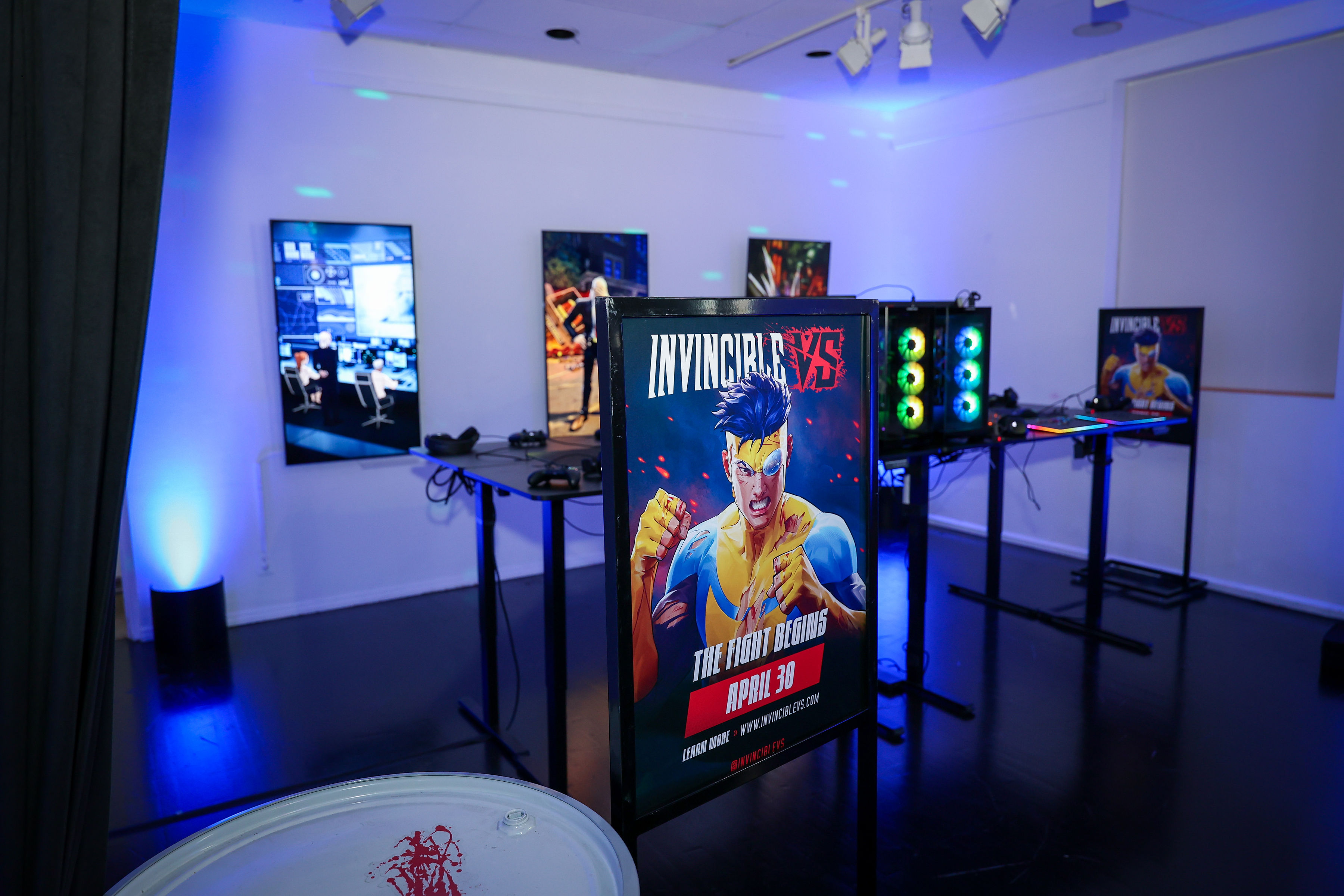 Dimly lit room with Invincible-branded gaming stations, RGB PC towers, and a freestanding Invincible VS poster of the hero in a yellow-and-blue suit, set under blue uplighting and glossy floors.