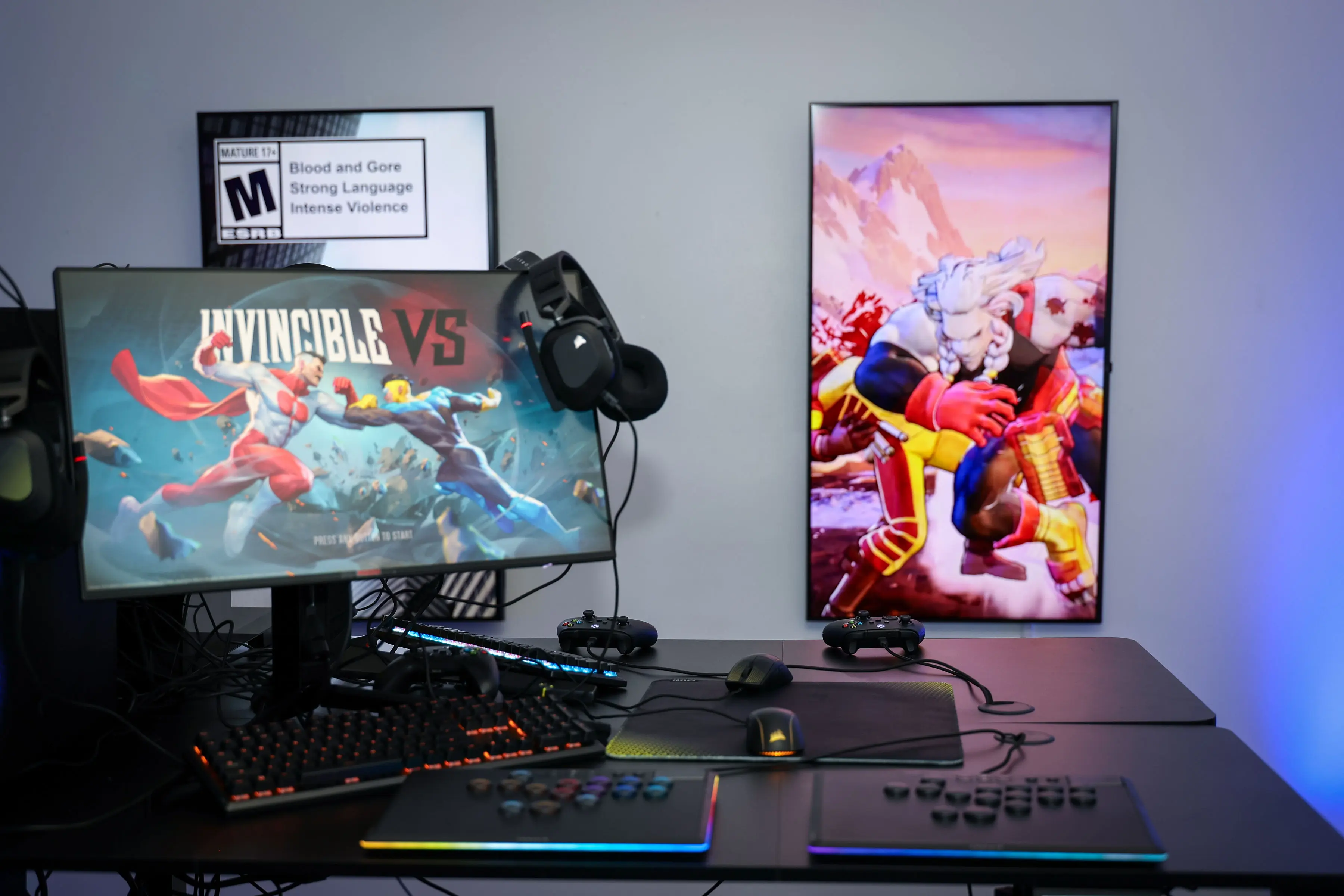 Gaming desk with RGB keyboard, mouse, controllers, and a widescreen monitor displaying 'INVINCIBLE VS' title art, hanging headphones, and a vertical character panel mounted on a neutral-colored wall.