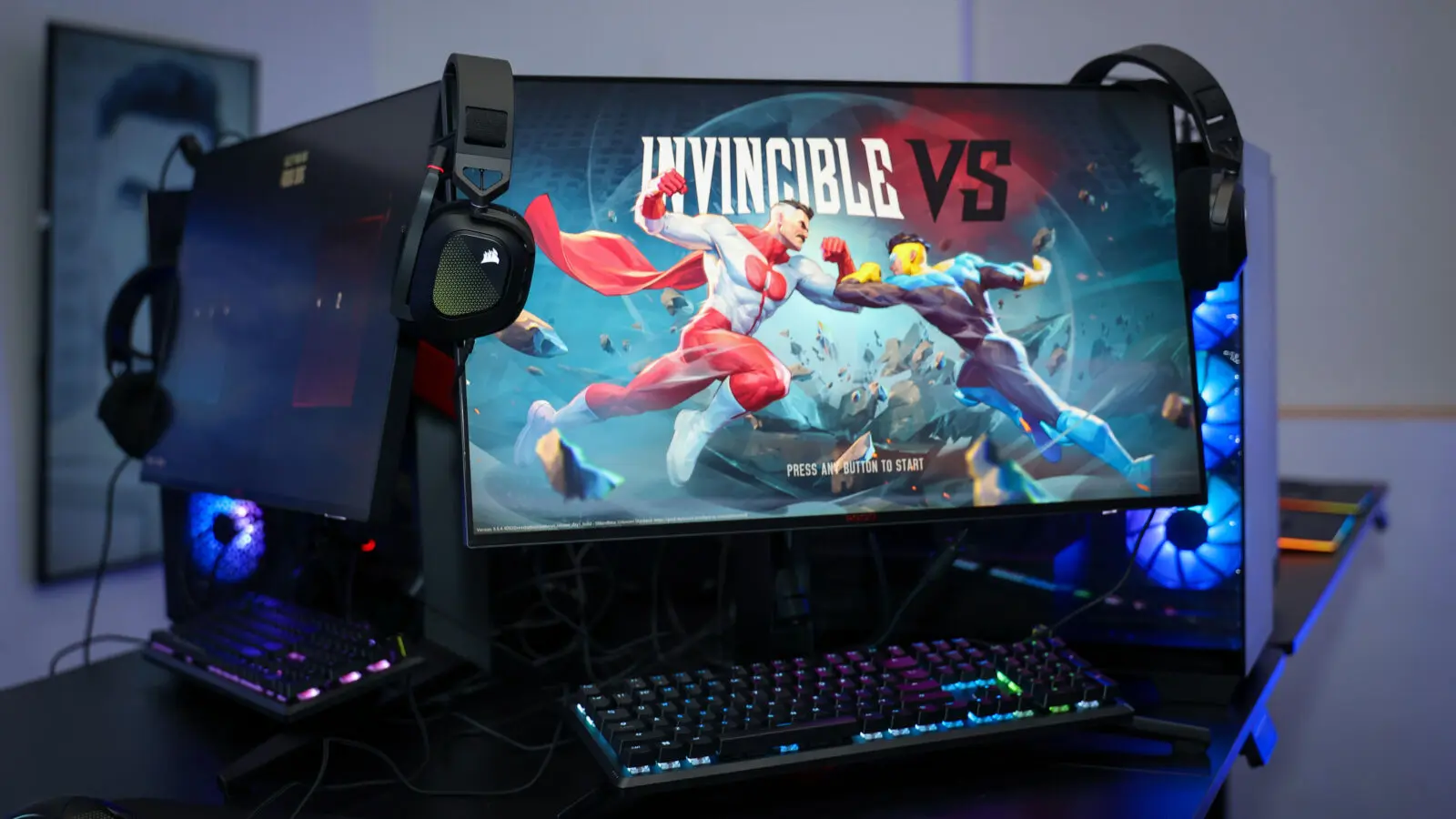 Ultra-wide monitor on a gaming station displays the 'INVINCIBLE VS' fight artwork between two superheroes, flanked by RGB-lit PC towers, keyboards, headsets, and mice on a black table.