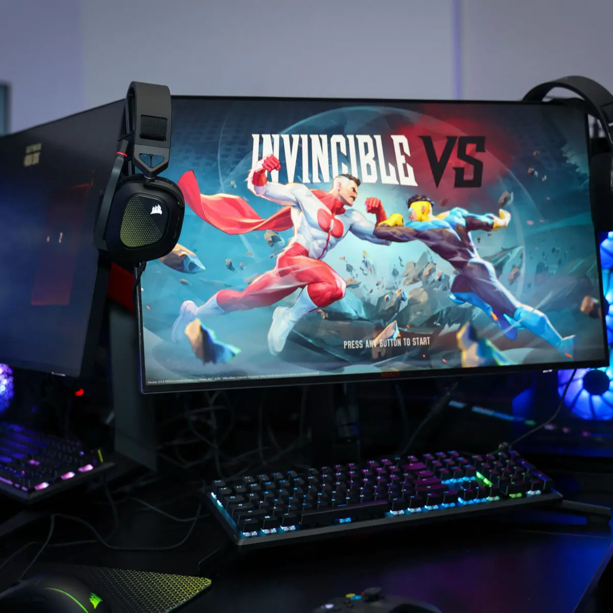 Ultra-wide monitor on a gaming station displays the 'INVINCIBLE VS' fight artwork between two superheroes, flanked by RGB-lit PC towers, keyboards, headsets, and mice on a black table.