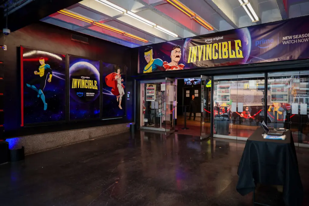 Theater lobby entrance decorated for Invincible Season 4 with three large character panels set against a space background, a wide INVINCIBLE banner above glass doors, and a check-in table.