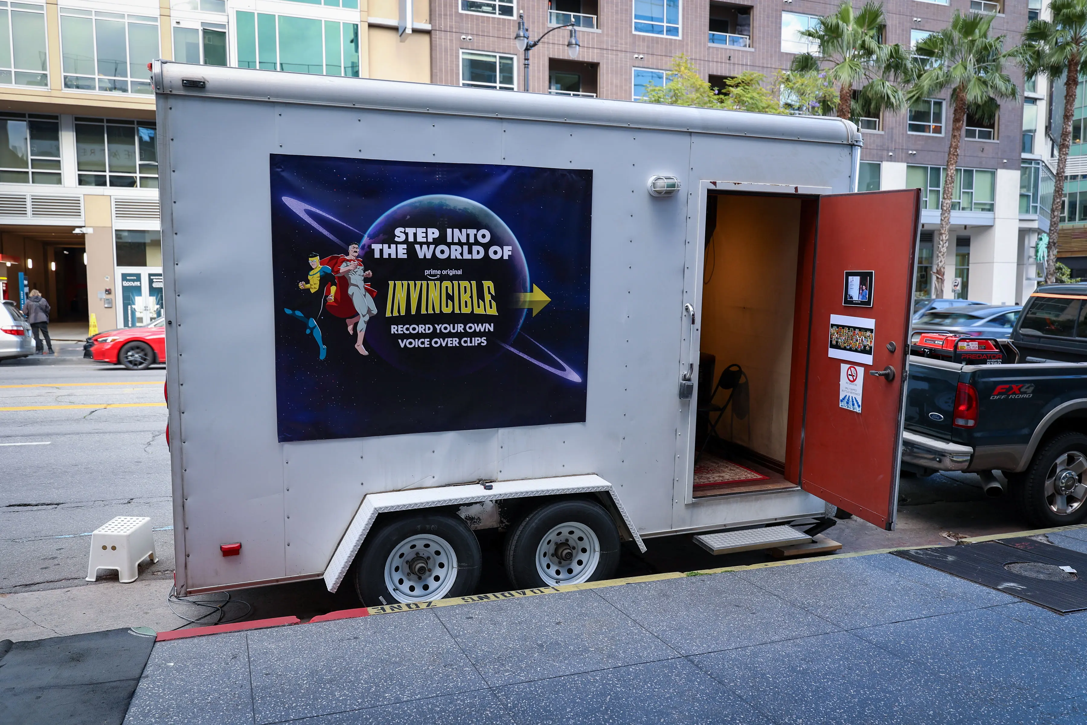 White mobile trailer parked on a sidewalk with an open red door revealing a small interior chair and rug, and a large Invincible poster that reads 'Record your own voice over clips'.