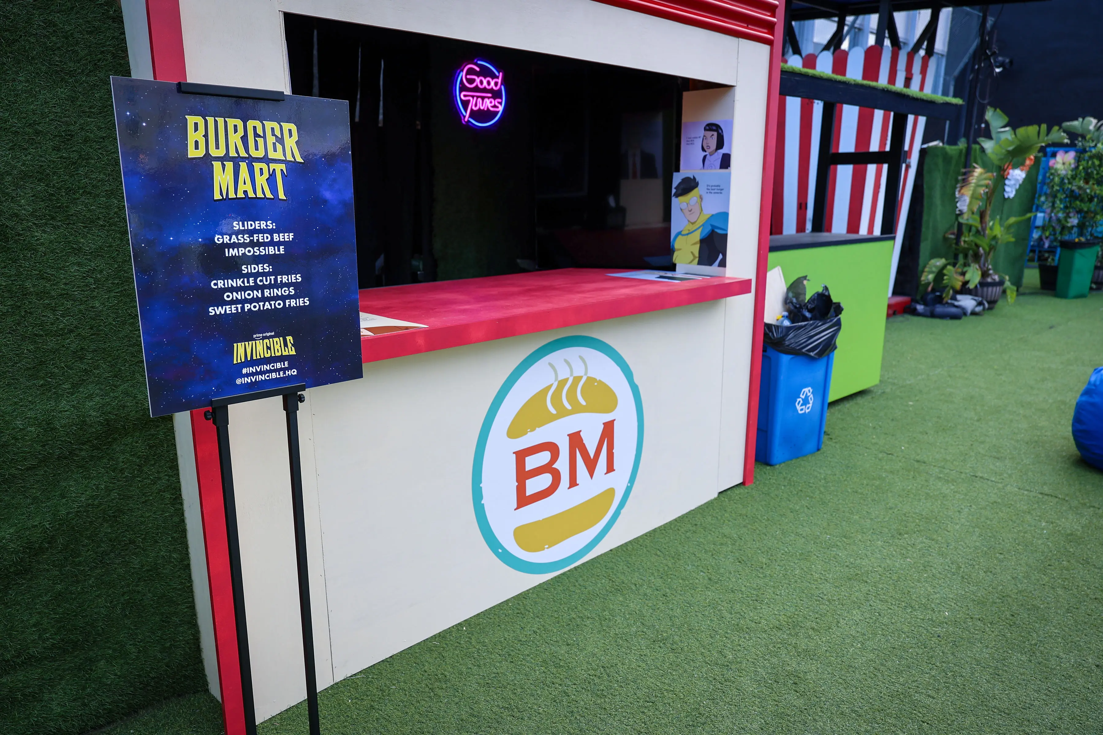Outdoor branded Burger Mart concession with red countertop and circular BM logo, a freestanding menu sign listing sliders and sides, a neon 'Good Vibes' sign, and artificial turf floor.
