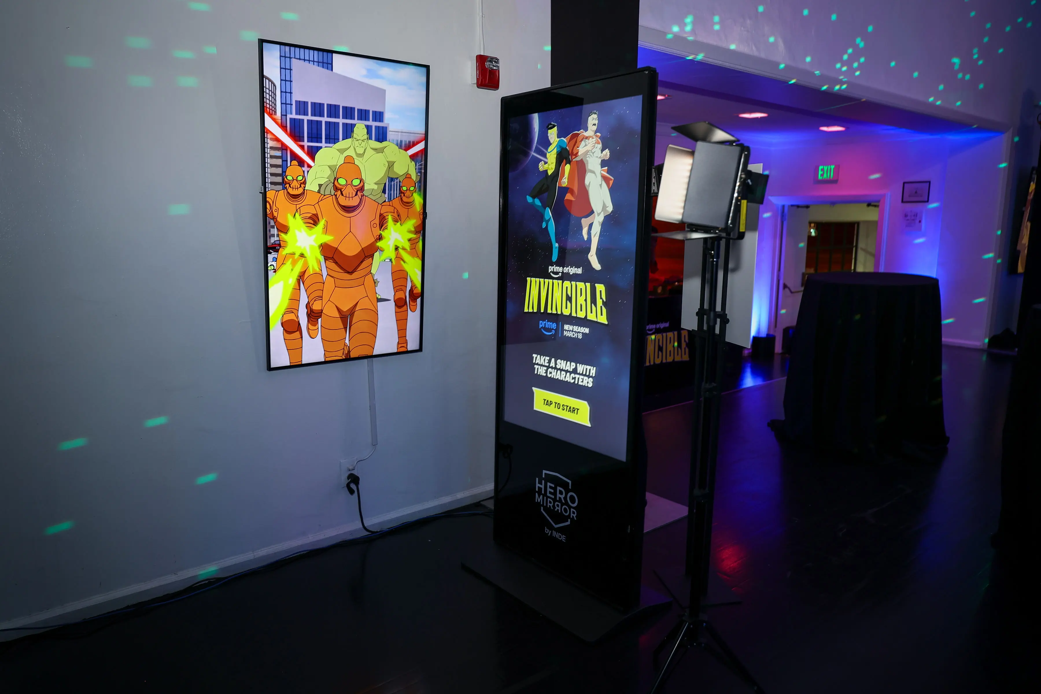 Freestanding touchscreen kiosk with Invincible hero artwork faces a wall-mounted animated panel showing armored enemies; studio lights and colored uplighting frame a phone-friendly creator photo area.