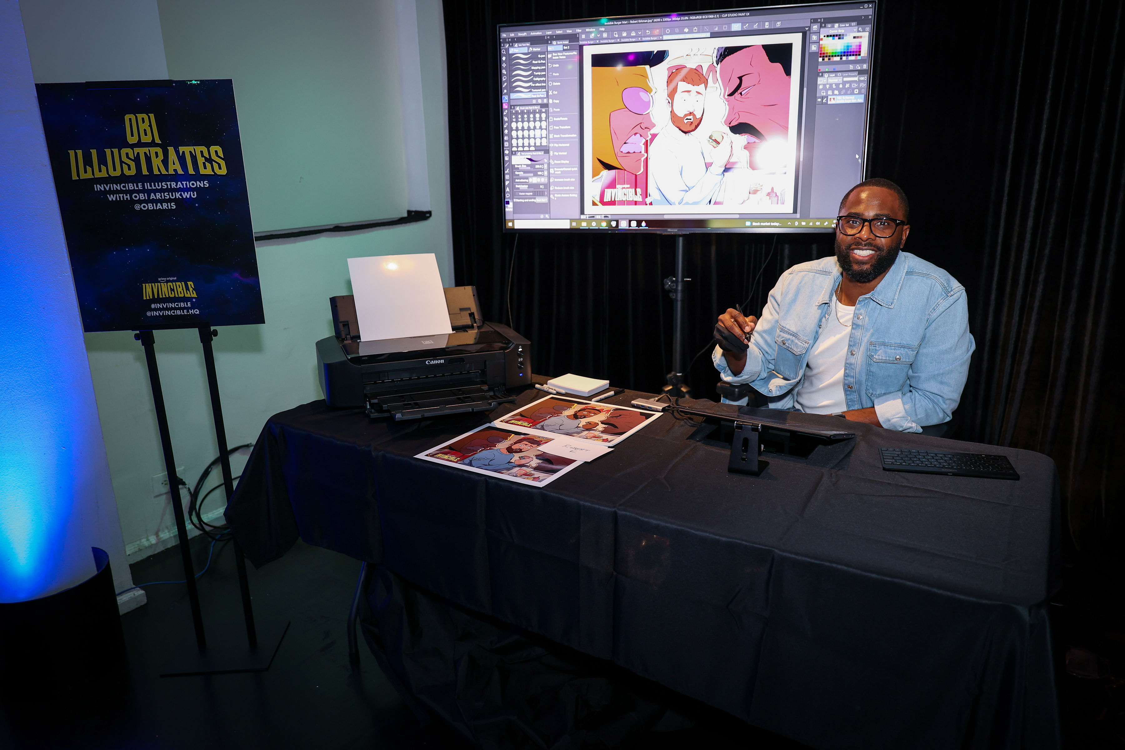 An illustrator seated at a table works on a drawing tablet while printed Invincible illustrations, a Canon printer, and a monitor showing live digital artwork anchor a Season 4 creator station.