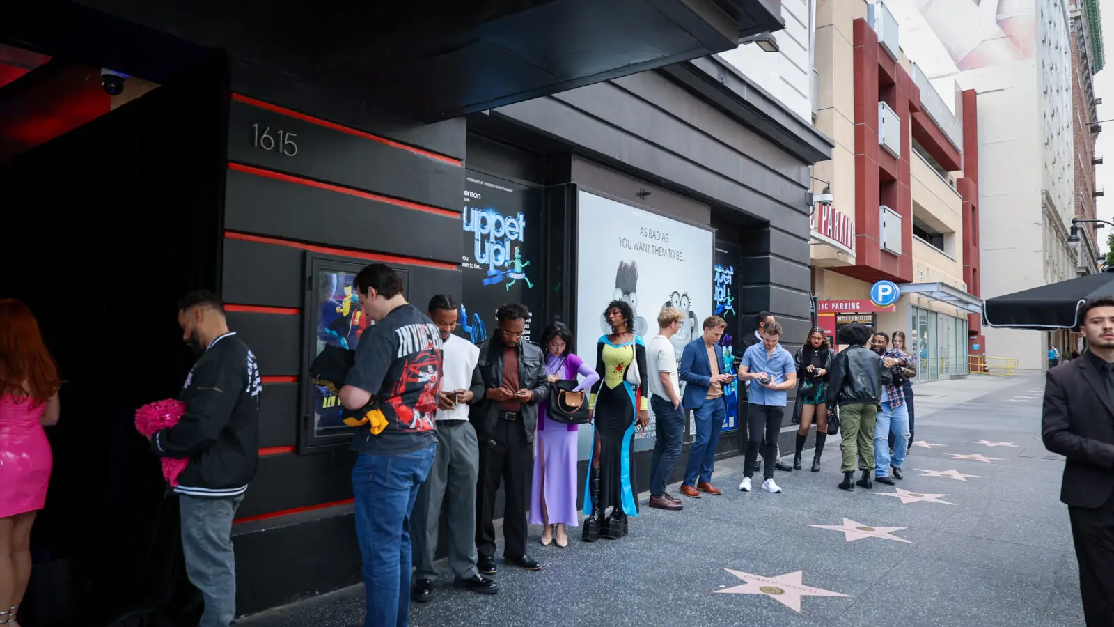 Line of attendees and creators in varied outfits waiting outside a screening venue entrance on a city sidewalk, with branded posters on the wall and Hollywood Walk of Fame stars underfoot.