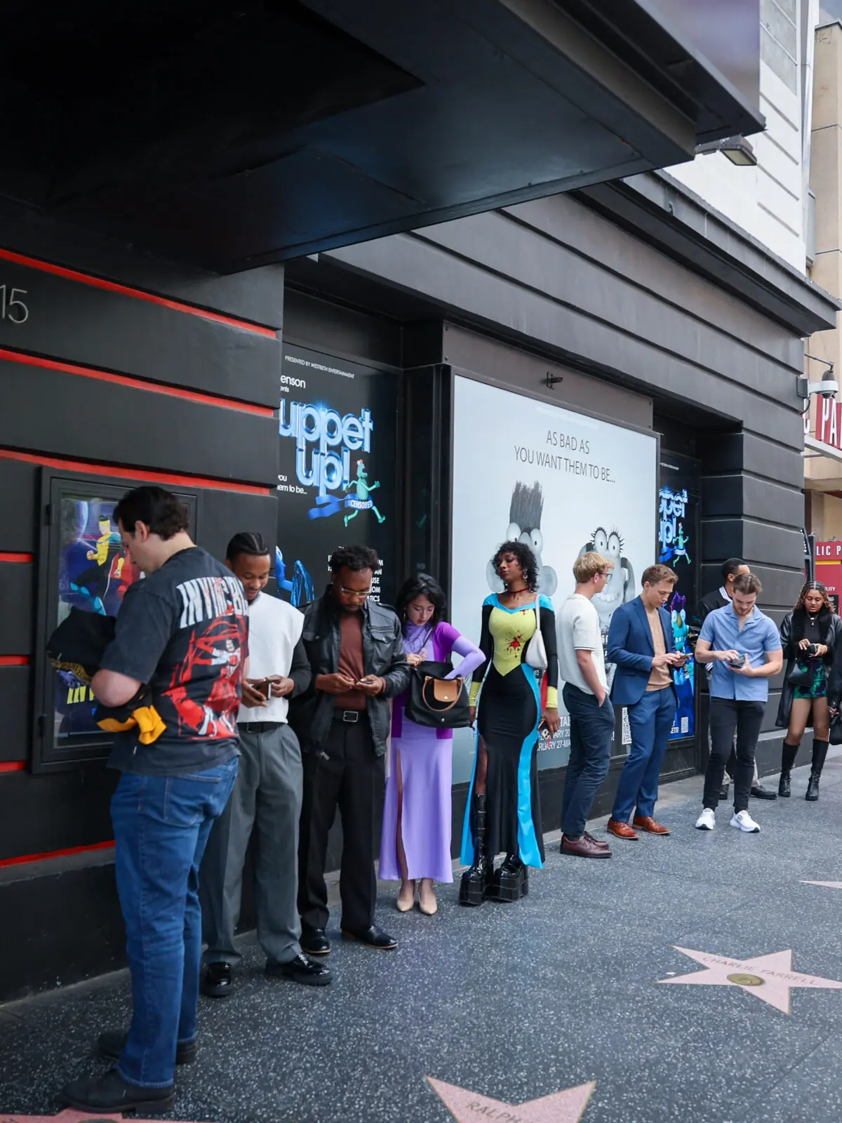 Line of attendees and creators in varied outfits waiting outside a screening venue entrance on a city sidewalk, with branded posters on the wall and Hollywood Walk of Fame stars underfoot.