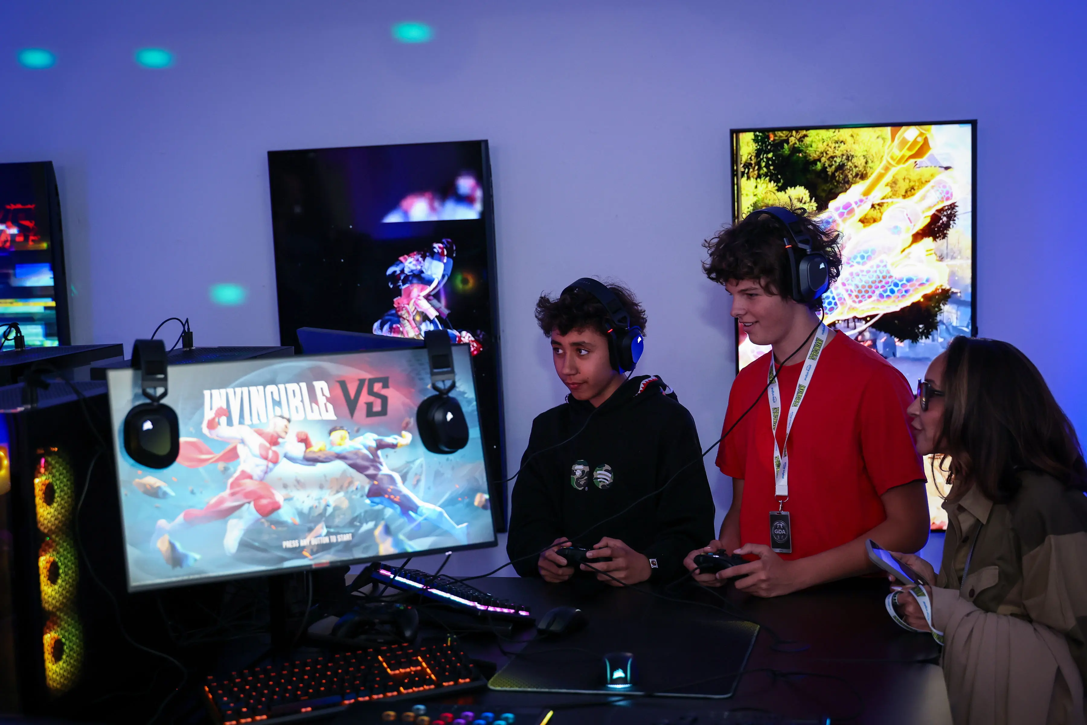 Three young creators wearing headphones play controllers at a branded gaming station with an INVINCIBLE VS match on the monitor, RGB PC fans glowing and vertical promo screens behind them.