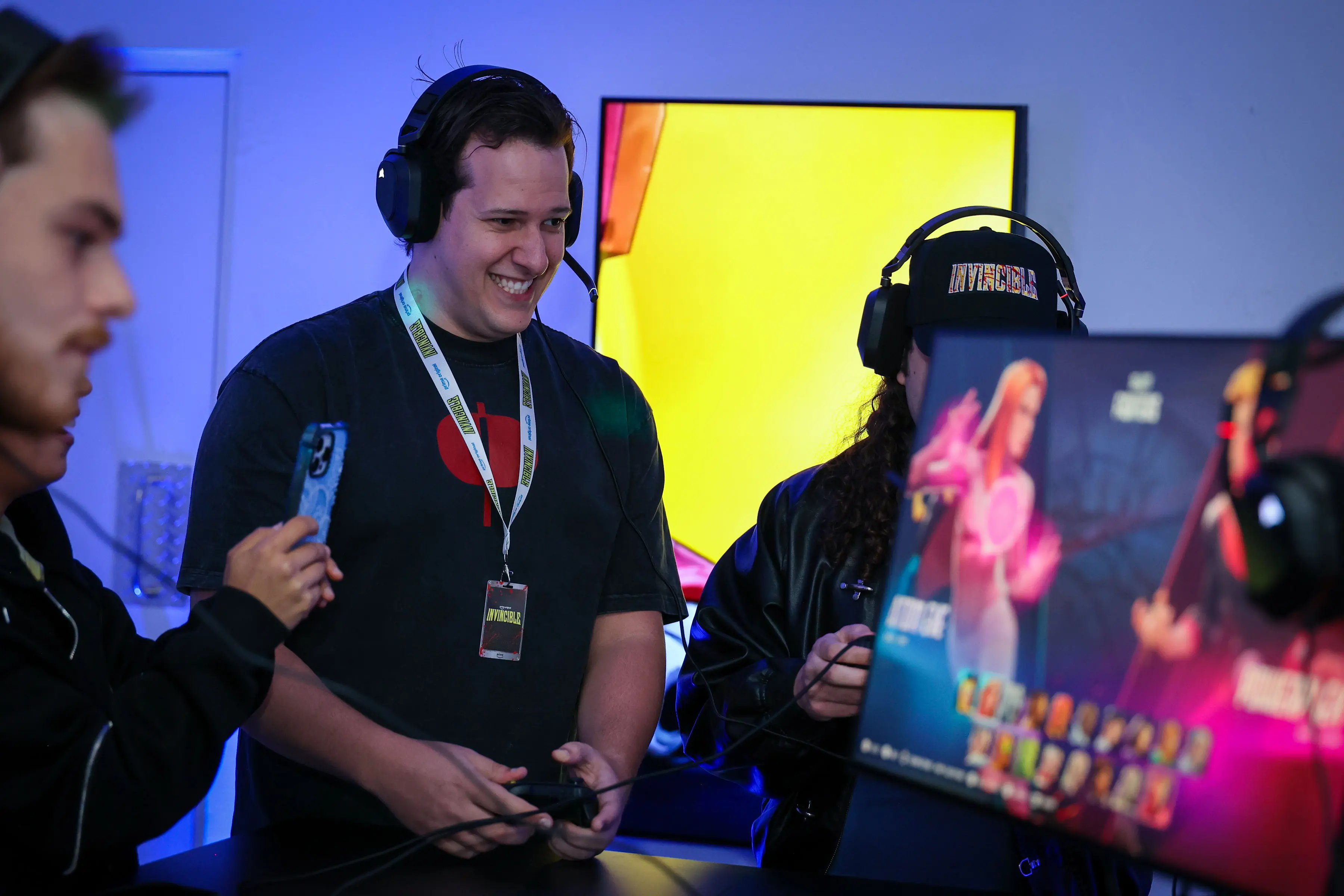 Attendees at a branded gaming station, one smiling with a headset and controller while another films on a phone; a monitor shows stylized character art against a bright yellow panel and cool blue gels.