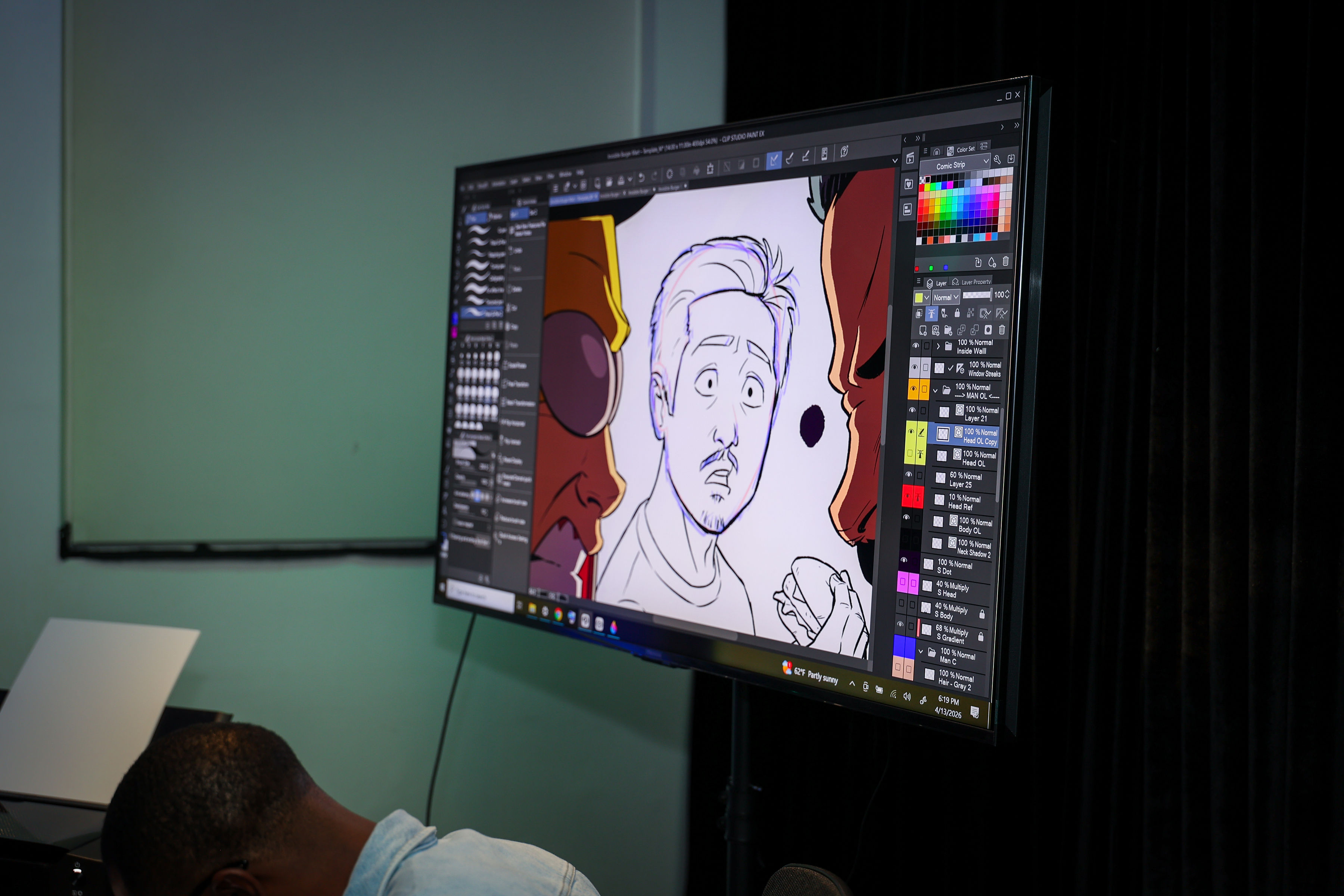 Curved monitor displaying a digital comic illustration of a surprised man flanked by two helmeted figures, with the drawing app interface visible and a person seated at the workstation foreground.