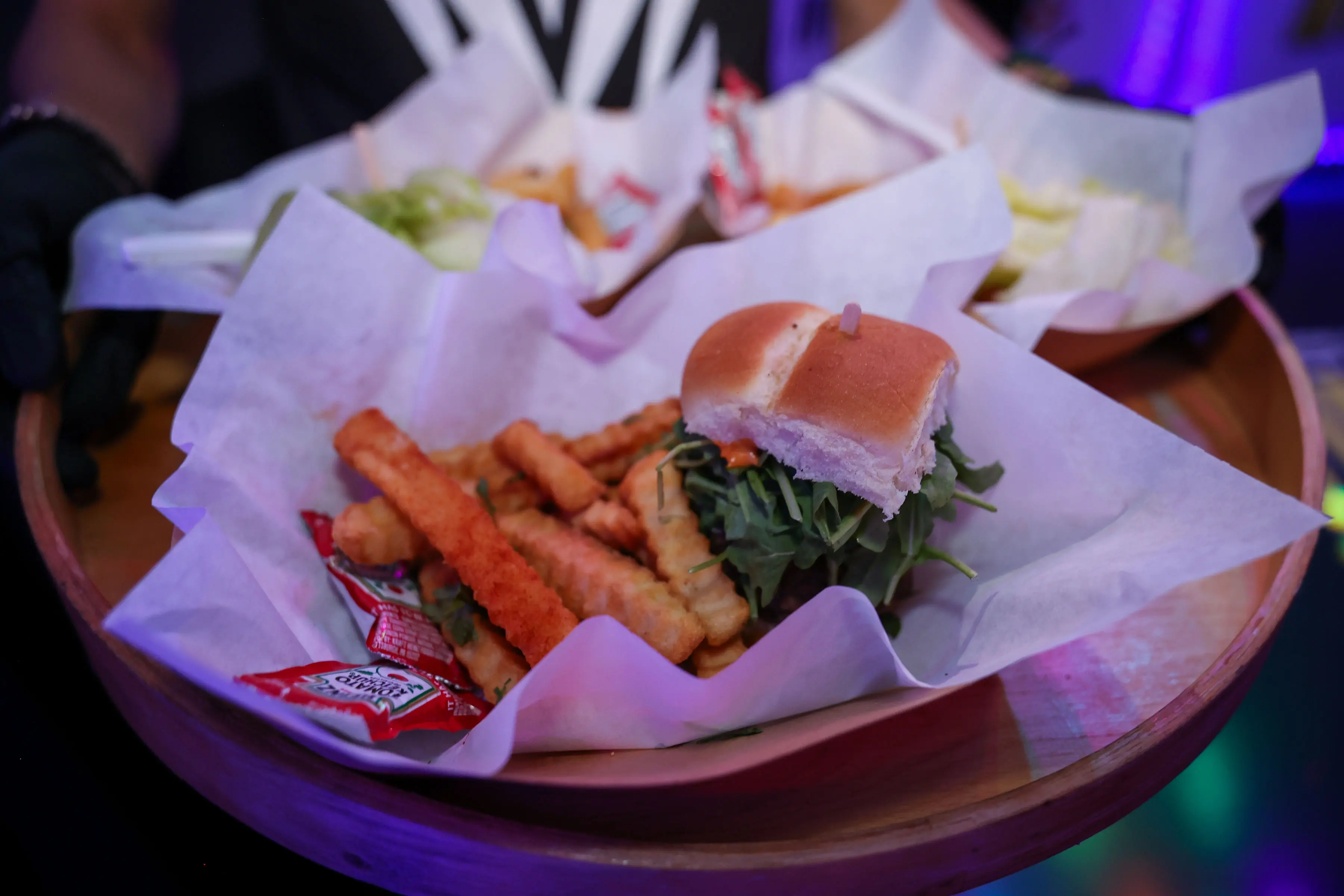 Tray with a small slider topped with leafy greens, crinkle-cut fries, and two ketchup packets on white deli paper, presented on a round wooden serving platter under purple event lighting.