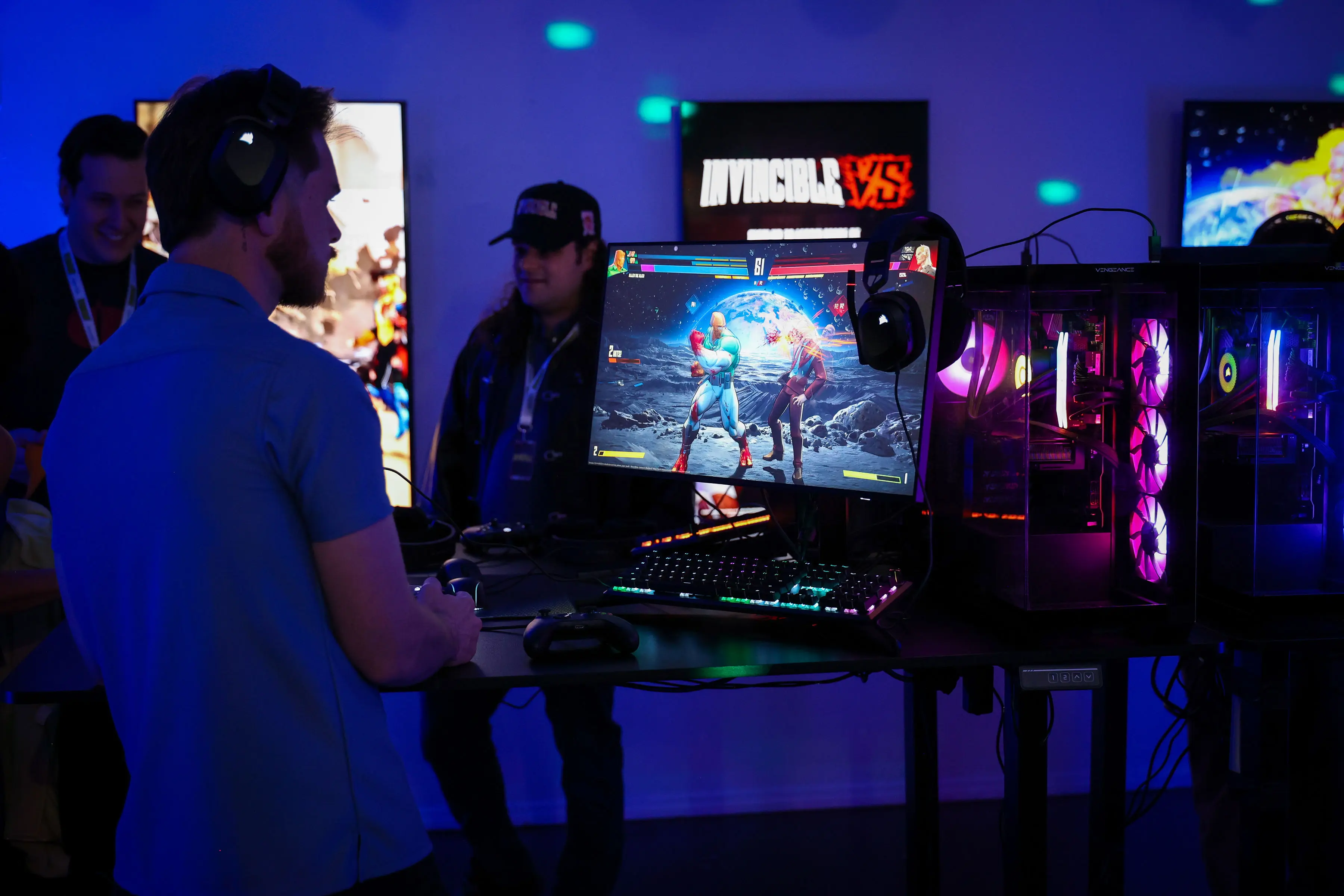 A headset-wearing attendee plays a fighting game at an RGB-lit gaming station with monitor and transparent PC towers, while INVINCIBLE VS signage and blue-purple event lighting glow in the background.