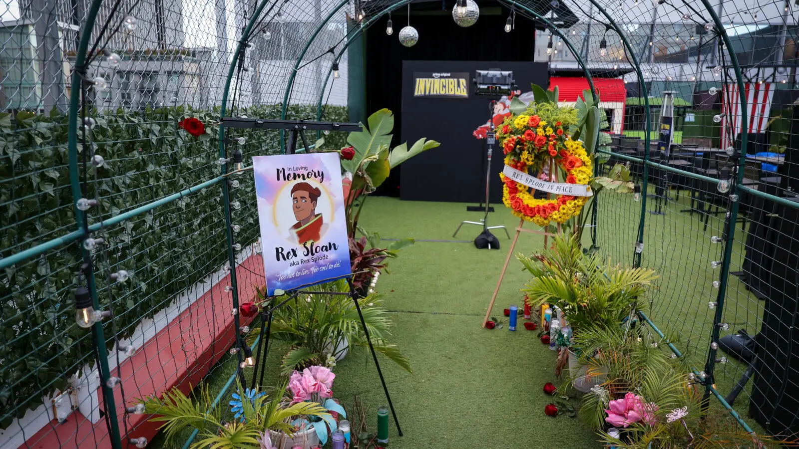 Arched green metal tunnel with string lights leading to an Invincible backdrop, flanked by a memorial poster for Rex Sloan (Rex Splode), a floral wreath, candles and potted plants on turf.