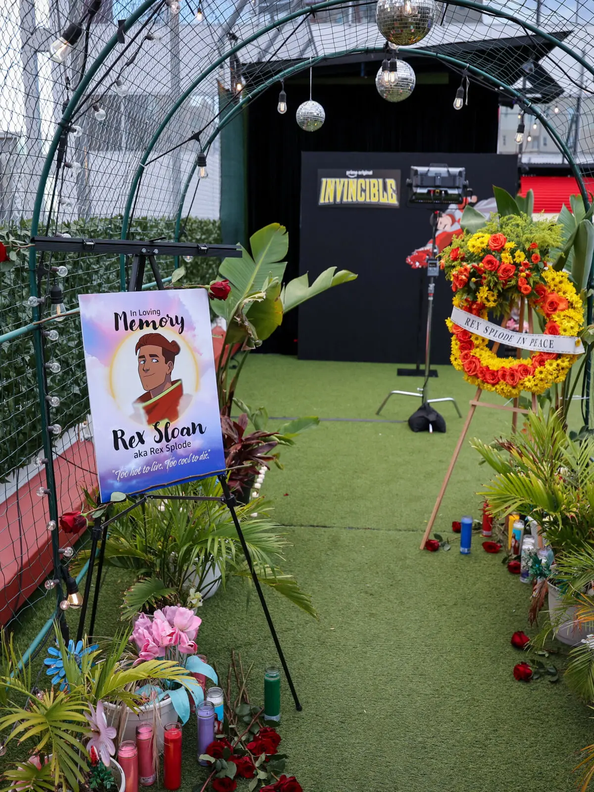 Arched green metal tunnel with string lights leading to an Invincible backdrop, flanked by a memorial poster for Rex Sloan (Rex Splode), a floral wreath, candles and potted plants on turf.