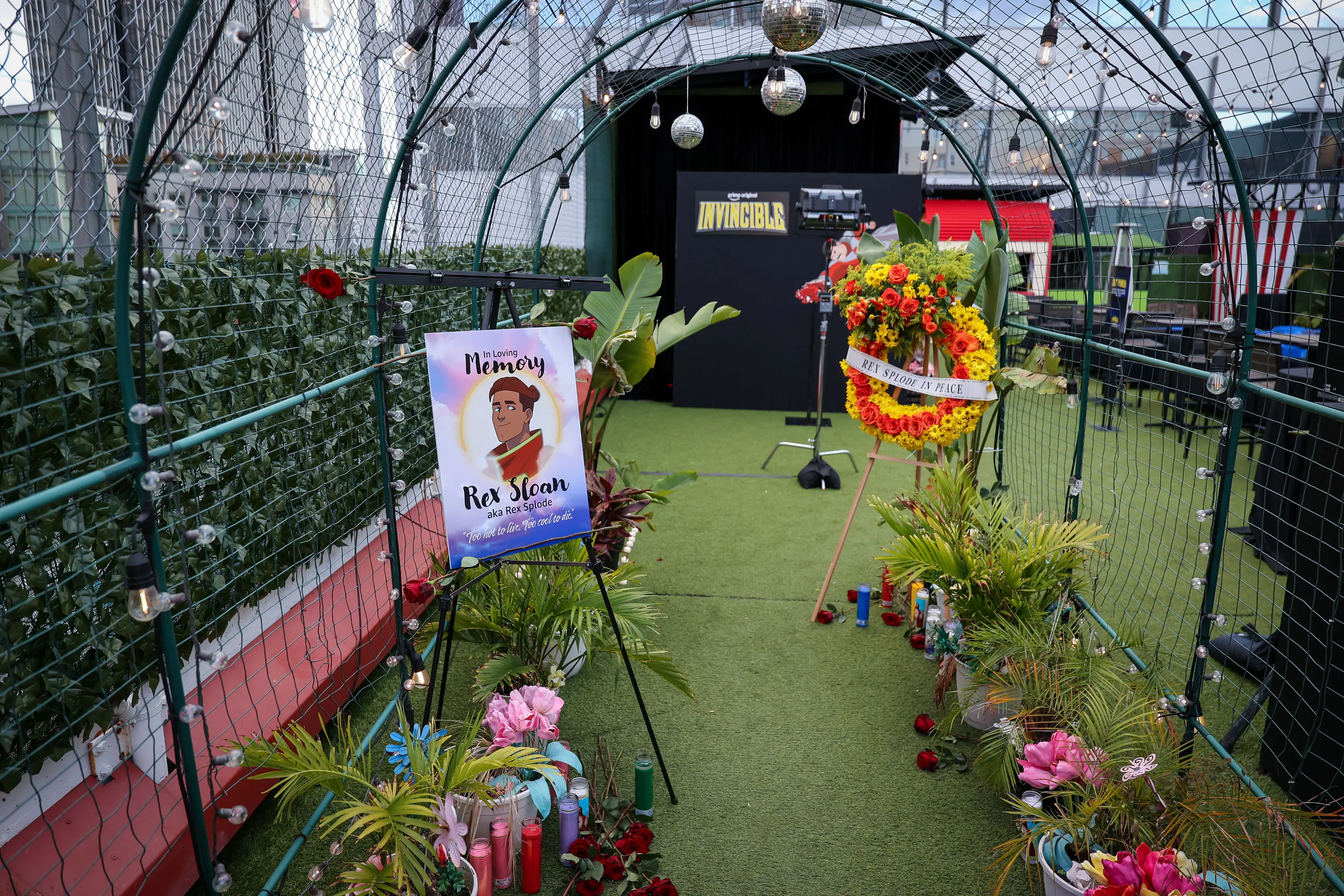 Arched memorial walkway on turf lined with candles, potted plants, a memorial easel reading 'Rex Sloan aka Rex Splode', a floral wreath, string lights, and an INVINCIBLE branded backdrop.