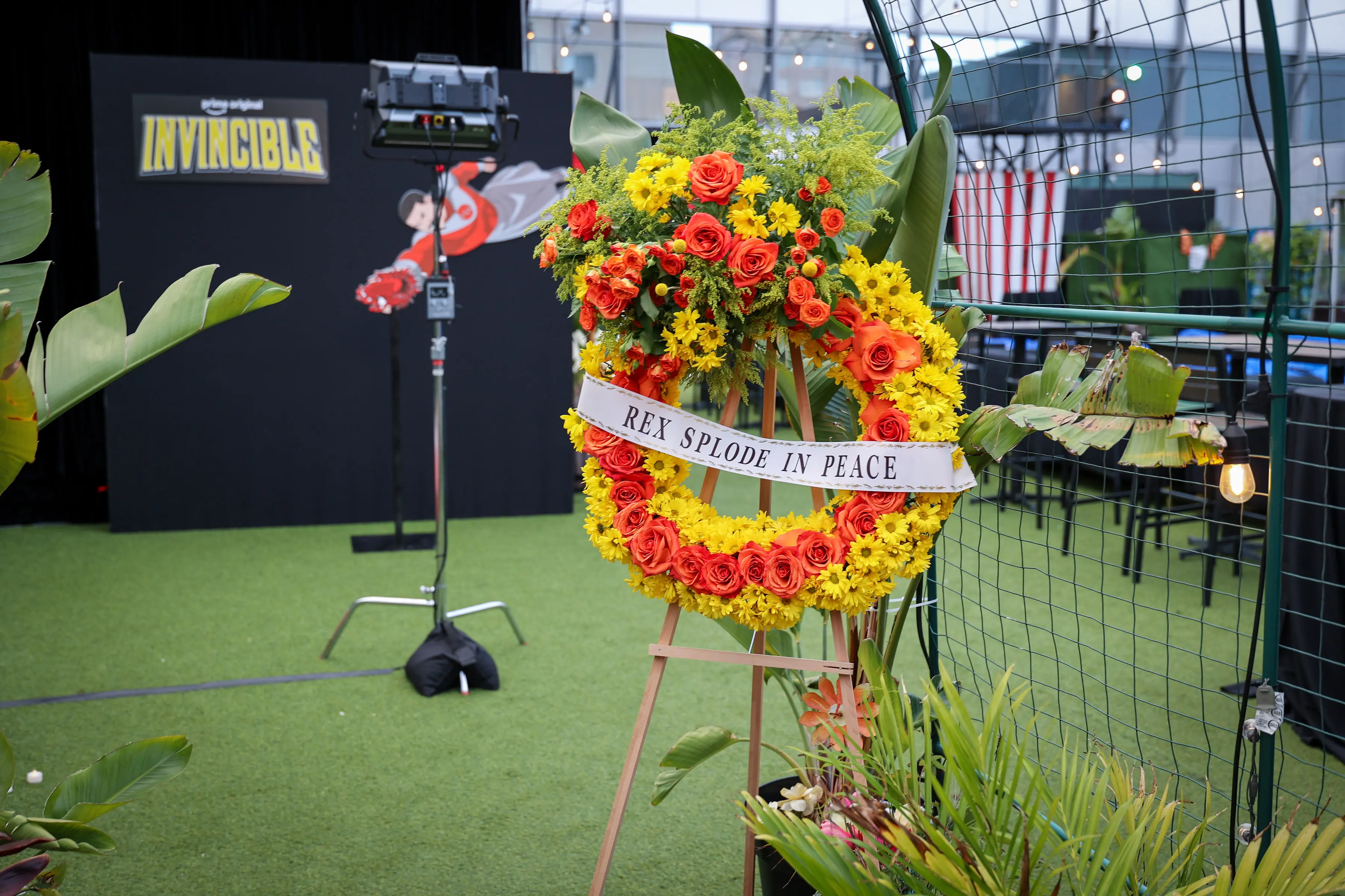Funeral-style wreath of orange roses and yellow daisies on an easel with a ribbon reading 'REX SPLODE IN PEACE,' staged in front of an Invincible logo backdrop.