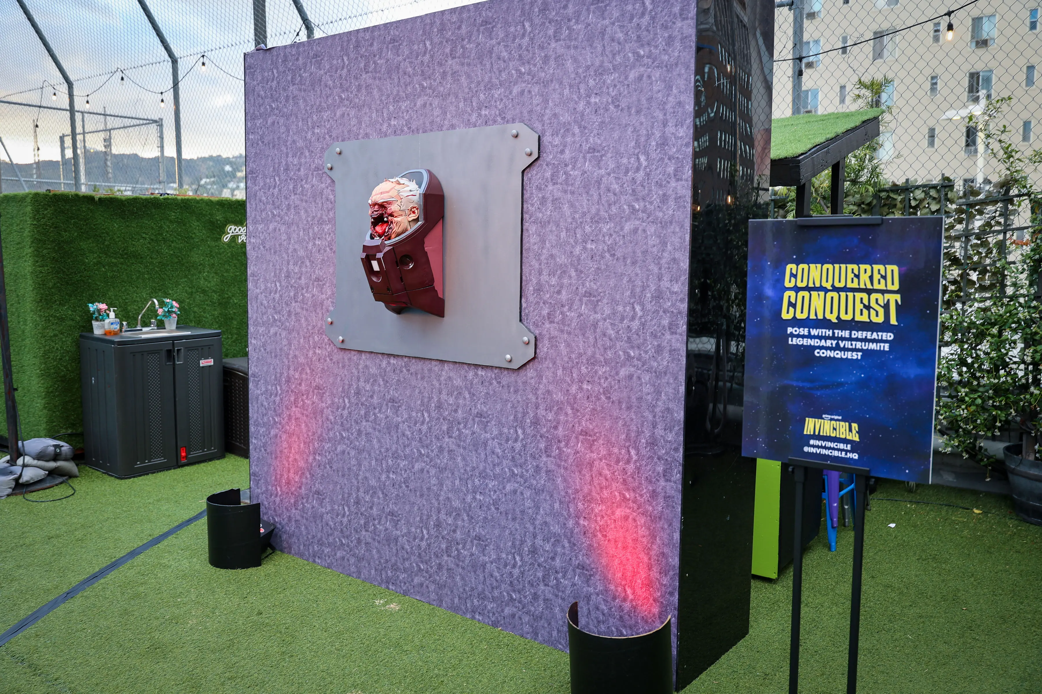 A blood-splattered prosthetic Viltrumite head mounted on a bolted metal plate attached to a purple textured wall, with red uplighting, turf flooring, and a nearby sign reading 'Conquered Conquest'.