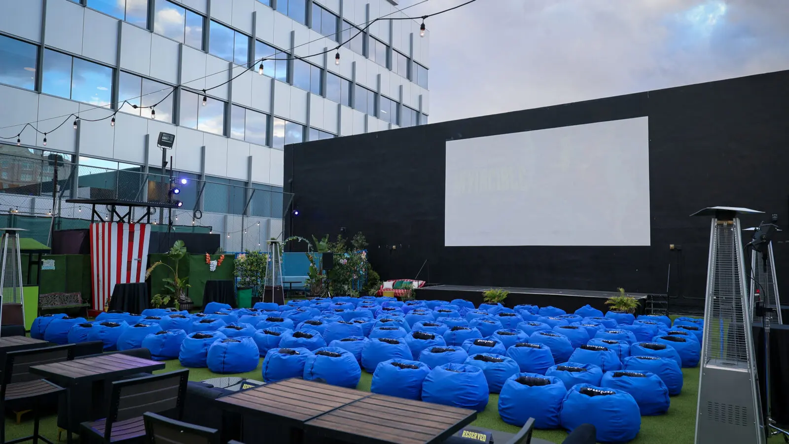 Rooftop screening setup with dozens of bright blue beanbag seats arranged facing a large white projection screen mounted on a black wall, surrounded by string lights, patio tables, plants, and heaters.
