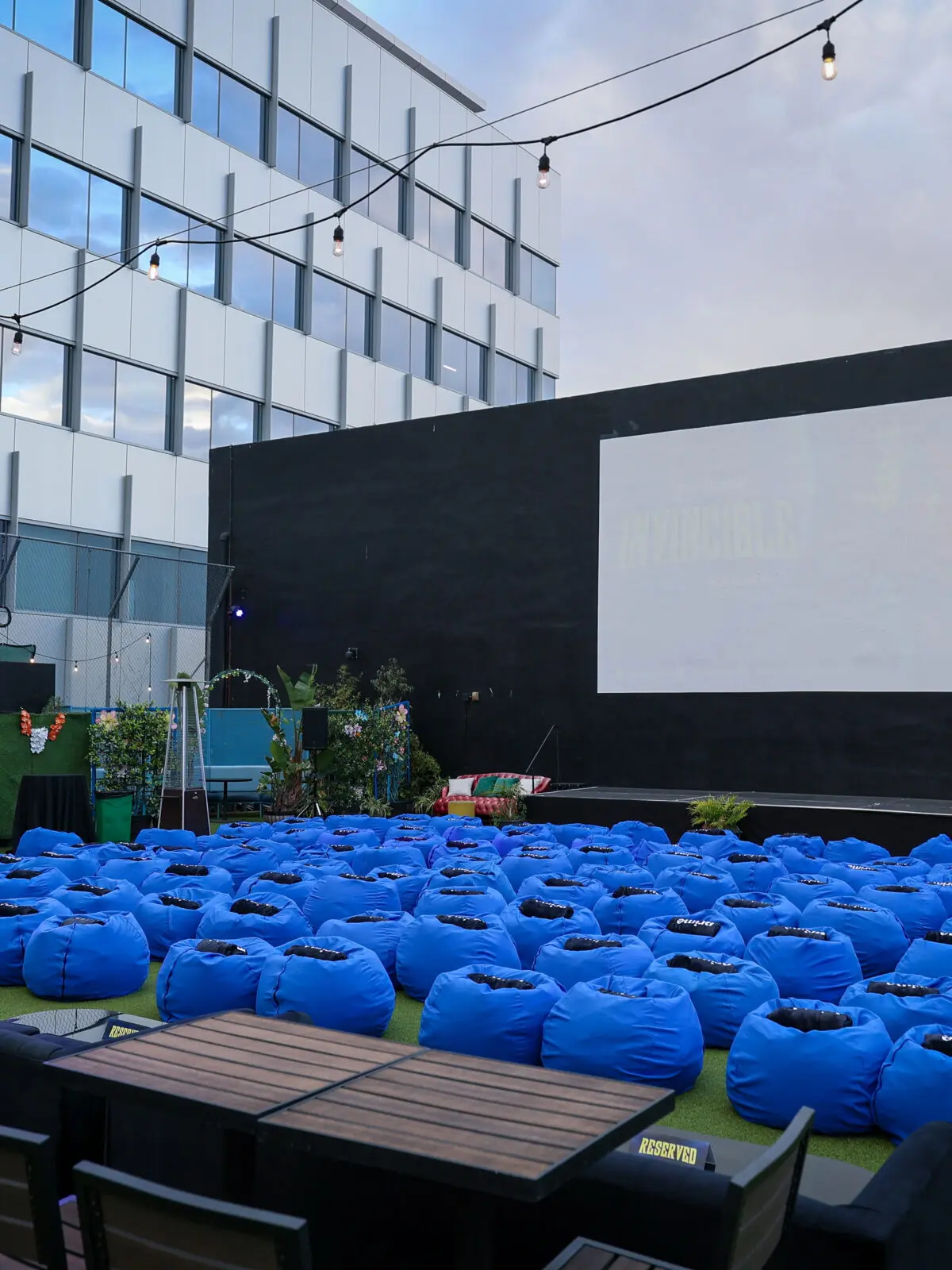 Rooftop screening setup with dozens of bright blue beanbag seats arranged facing a large white projection screen mounted on a black wall, surrounded by string lights, patio tables, plants, and heaters.