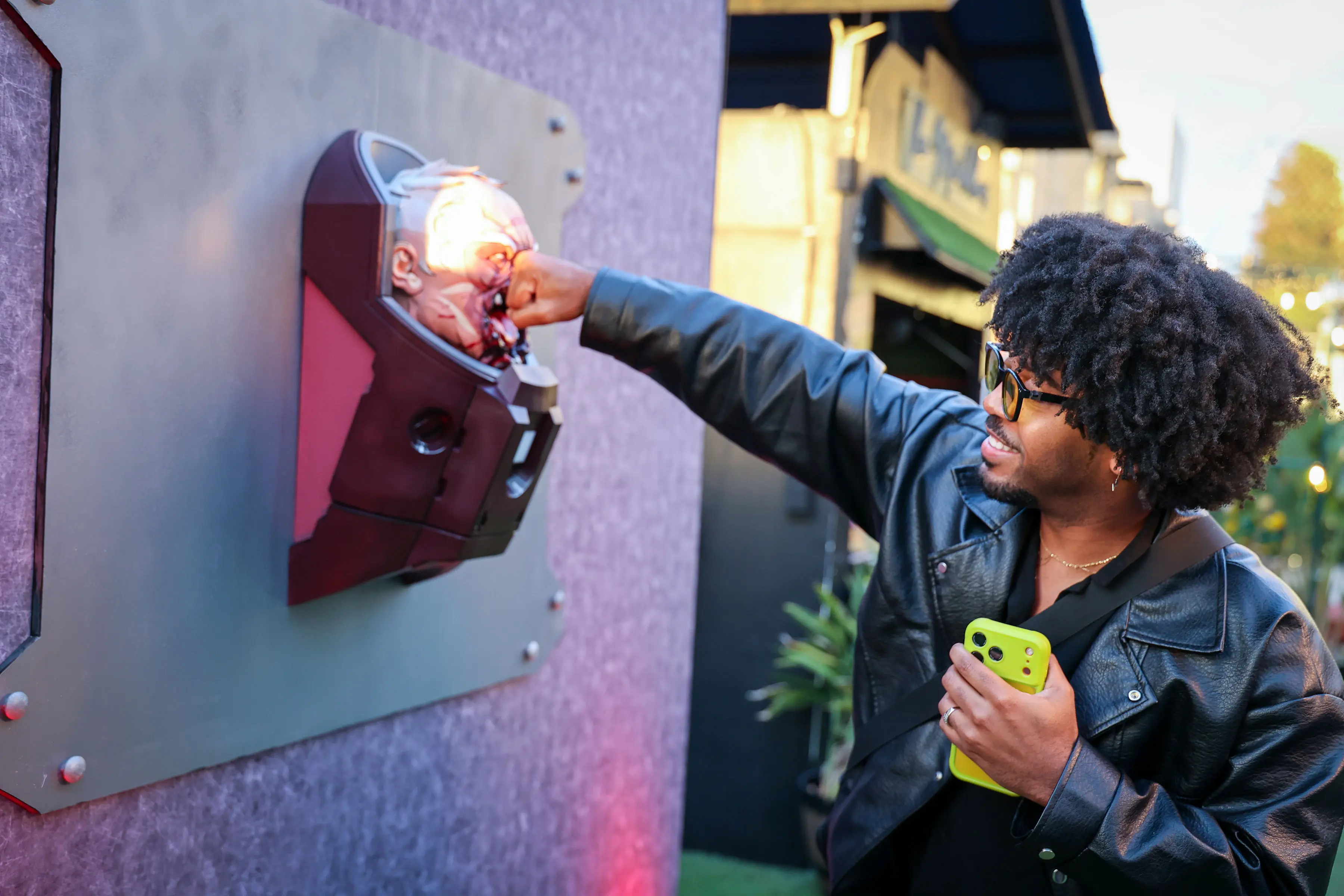 Attendee leaning into a blood-splattered prosthetic head mounted on a bolted panel while holding a neon-green phone, lit by bruised purple-red event lighting at an outdoor screening activation.