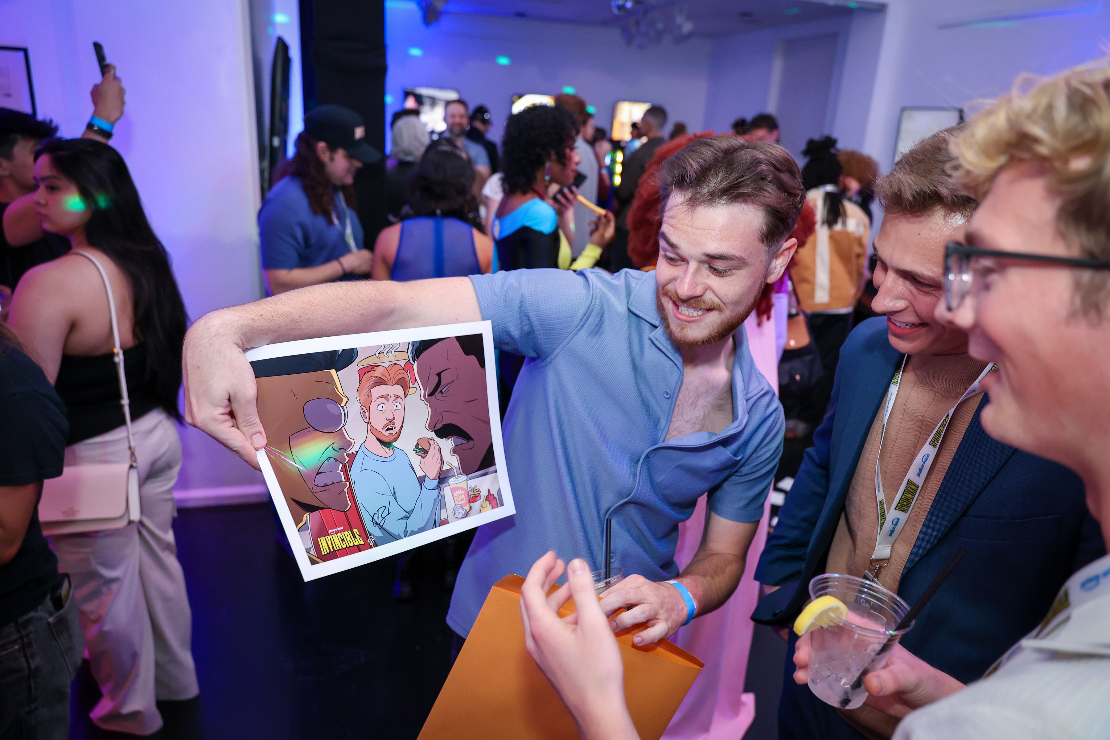 Three smiling guests in a crowded screening room examine a glossy Invincible illustration while blue and purple gel lighting and party guests blur in the background.