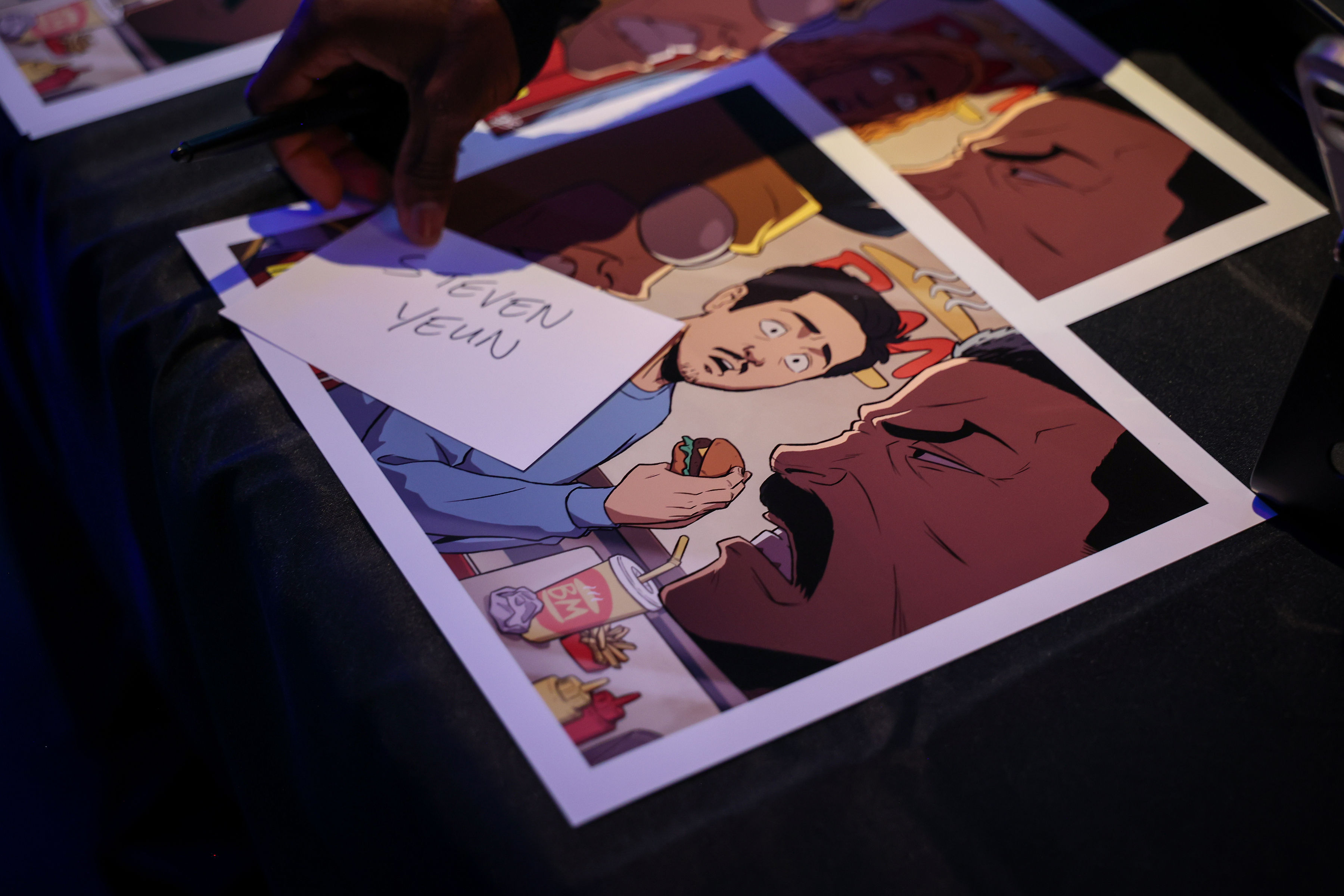 Handwritten 'STEVEN YEUN' guest card on top of a glossy comic-style illustration of two faces, sitting on a black-draped table under cool purple-blue event lighting.