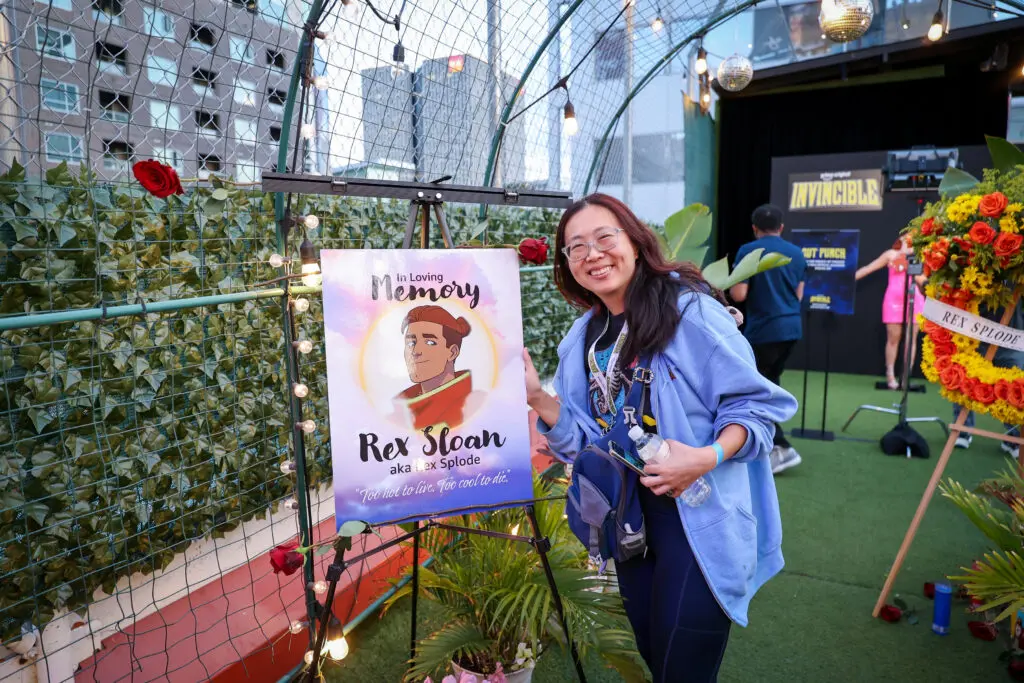 Smiling event attendee poses next to a lit memorial easel for Rex Splode with an illustrated portrait, floral wreath, and Invincible screening backdrop on a rooftop activation.
