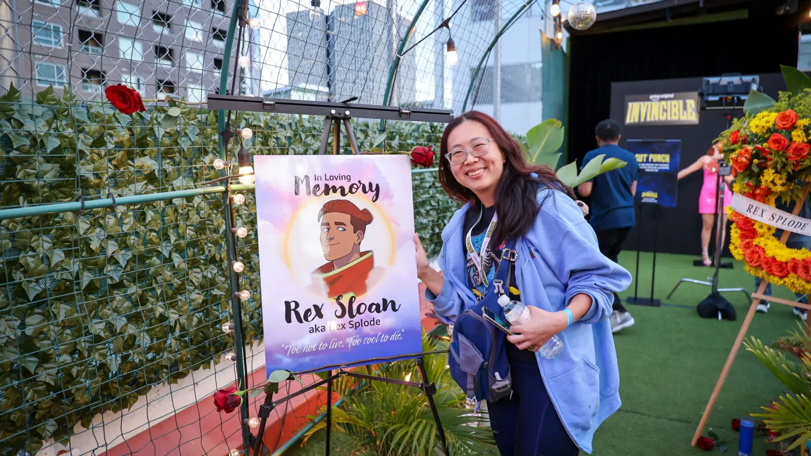 Smiling attendee in a blue hoodie poses next to an easel-mounted memorial poster reading "In Loving Memory Rex Sloan aka Rex Splode," surrounded by string lights, flowers, and Invincible branding in a rooftop screening.