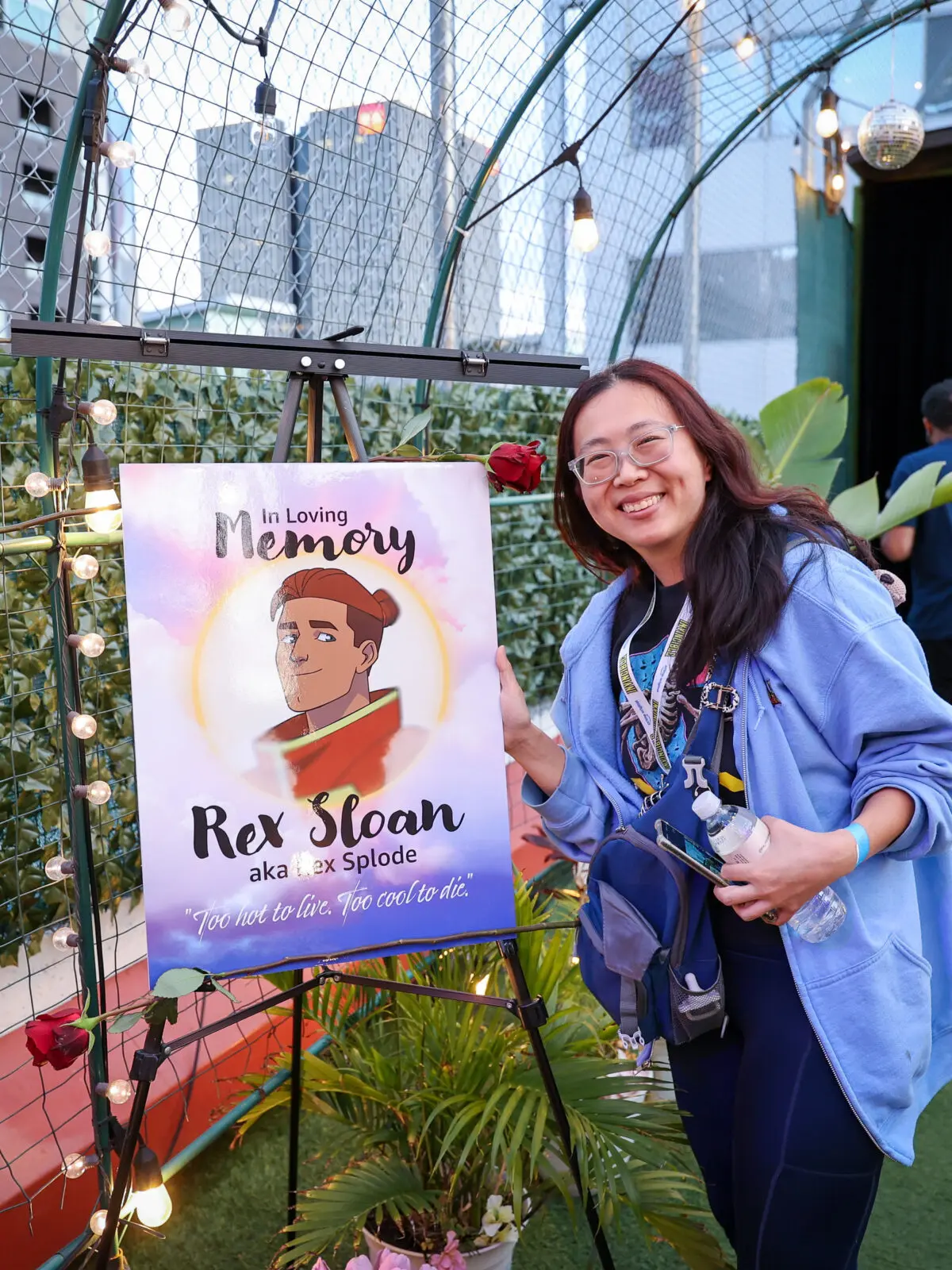 Smiling attendee in a blue hoodie poses next to an easel-mounted memorial poster reading "In Loving Memory Rex Sloan aka Rex Splode," surrounded by string lights, flowers, and Invincible branding in a rooftop screening.