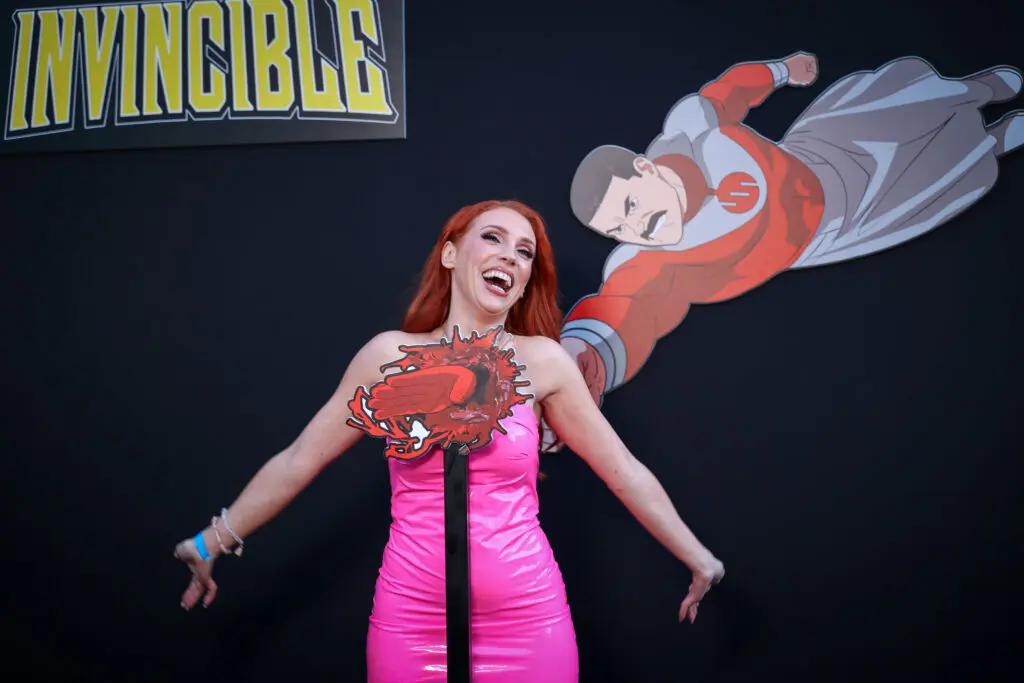 Creator Grace McClung poses beneath a flying illustrated Invincible cutout, holding a red comic-book impact prop at the Invincible Season 4 screening event.