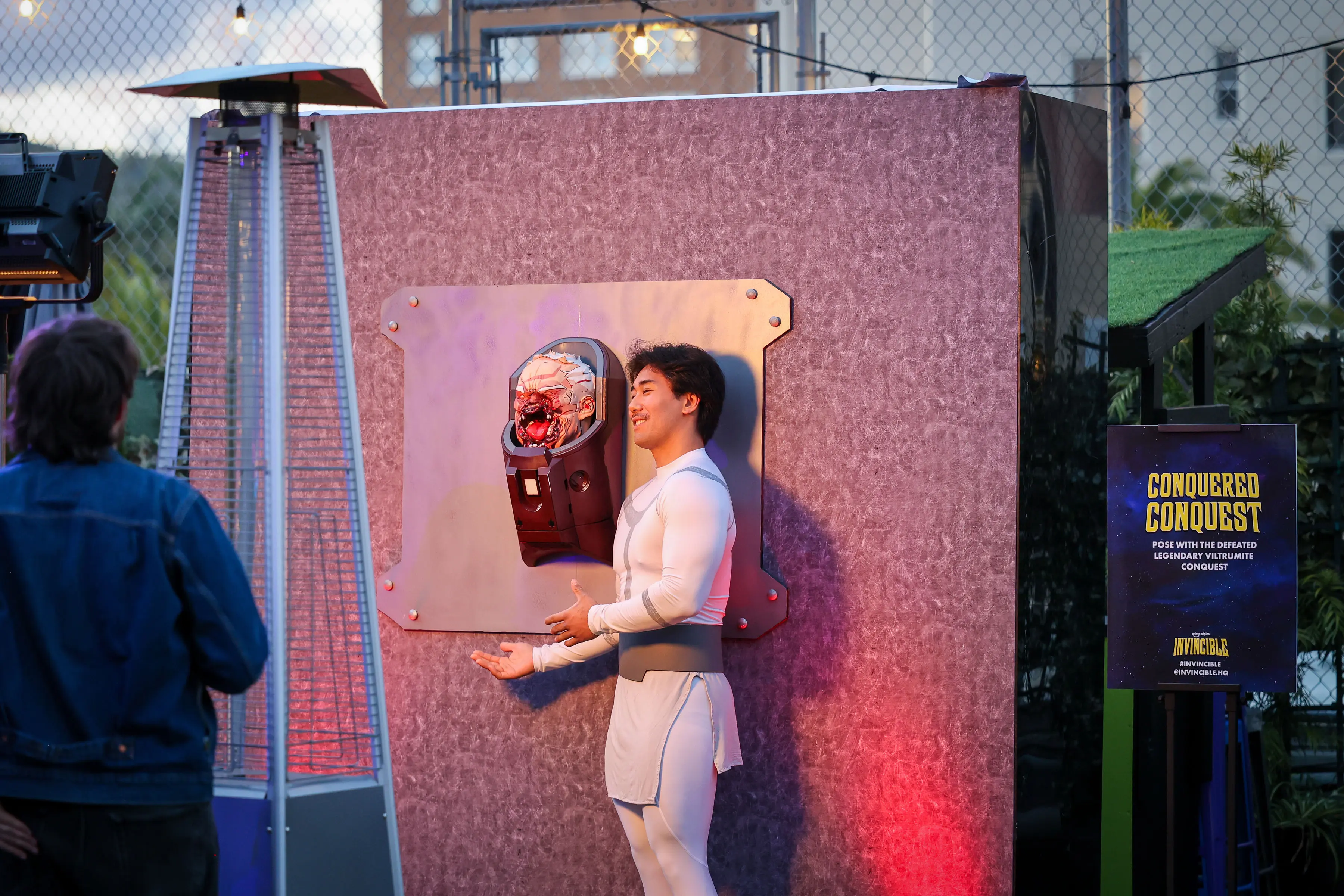A creator in a white costume poses beside a blood-splattered prosthetic head mounted on a bolted metal plate at an outdoor event with signage reading "Conquered Conquest" and colored event lighting.
