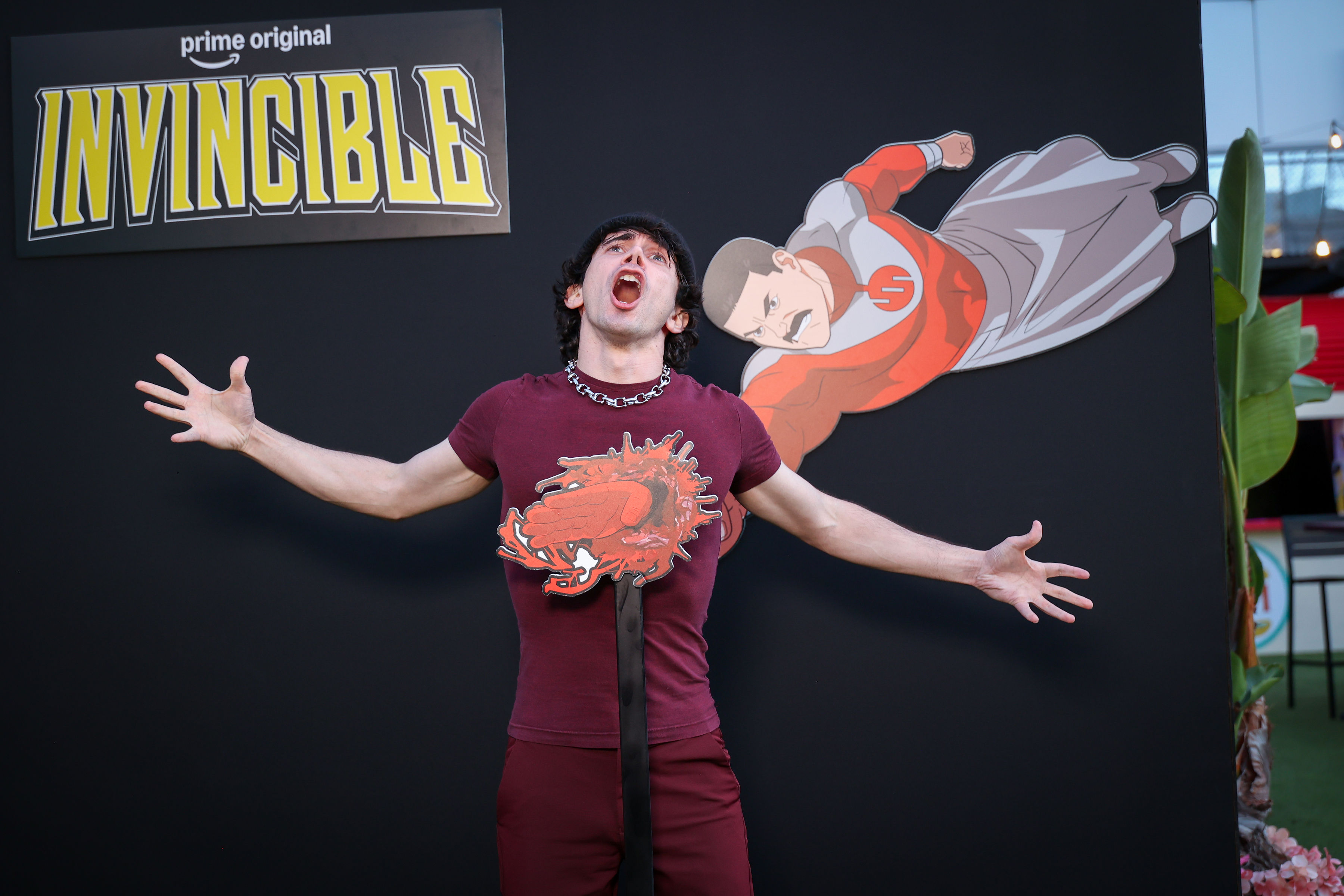 Guest in a maroon outfit stands with arms out and mouth open against a black backdrop bearing the yellow Invincible logo, next to a flying superhero cutout and a red chest-mounted prop.