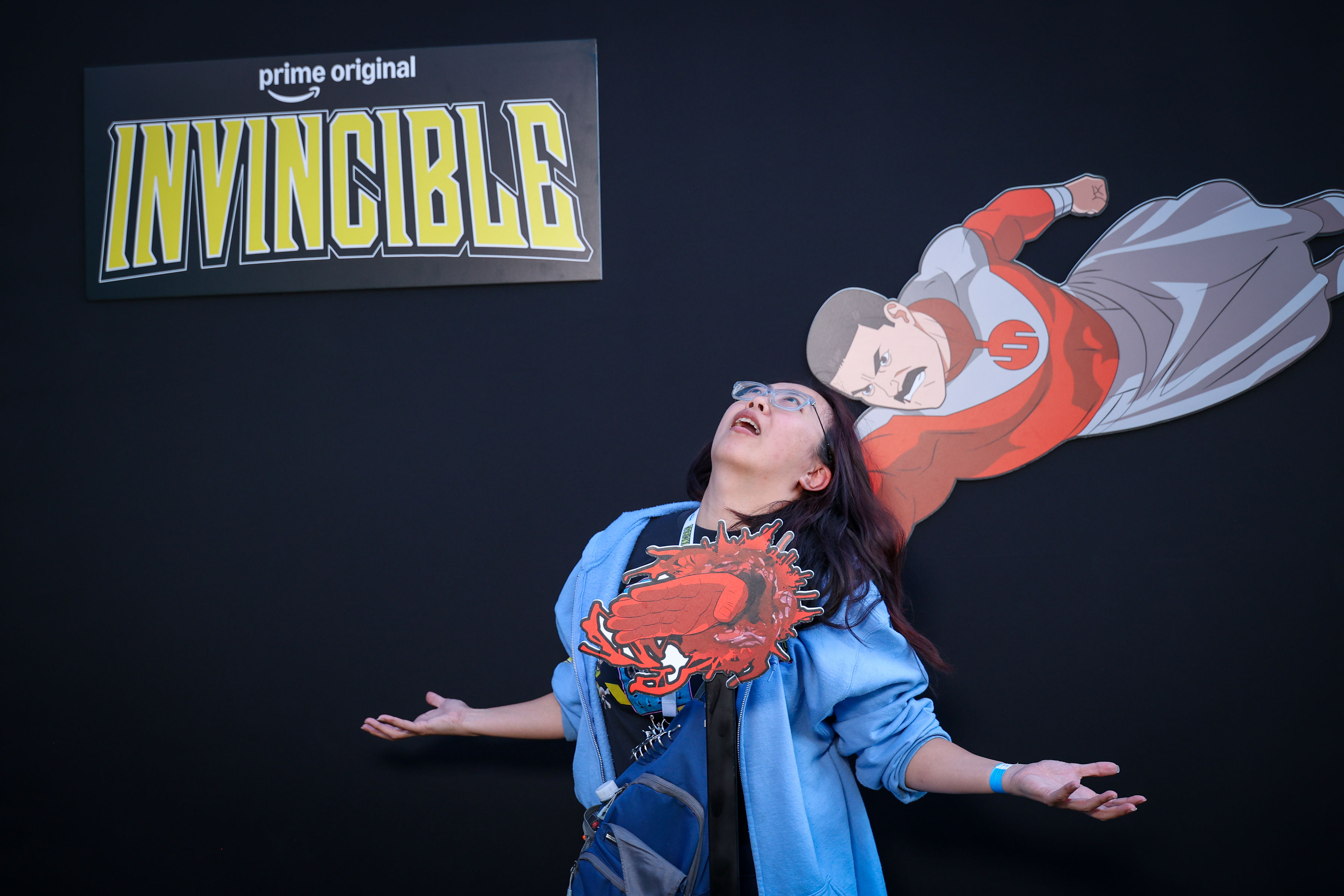 Person looks upward with arms outstretched next to an Omni-Man cardboard cutout and an 'Invincible' logo on a dark event wall.