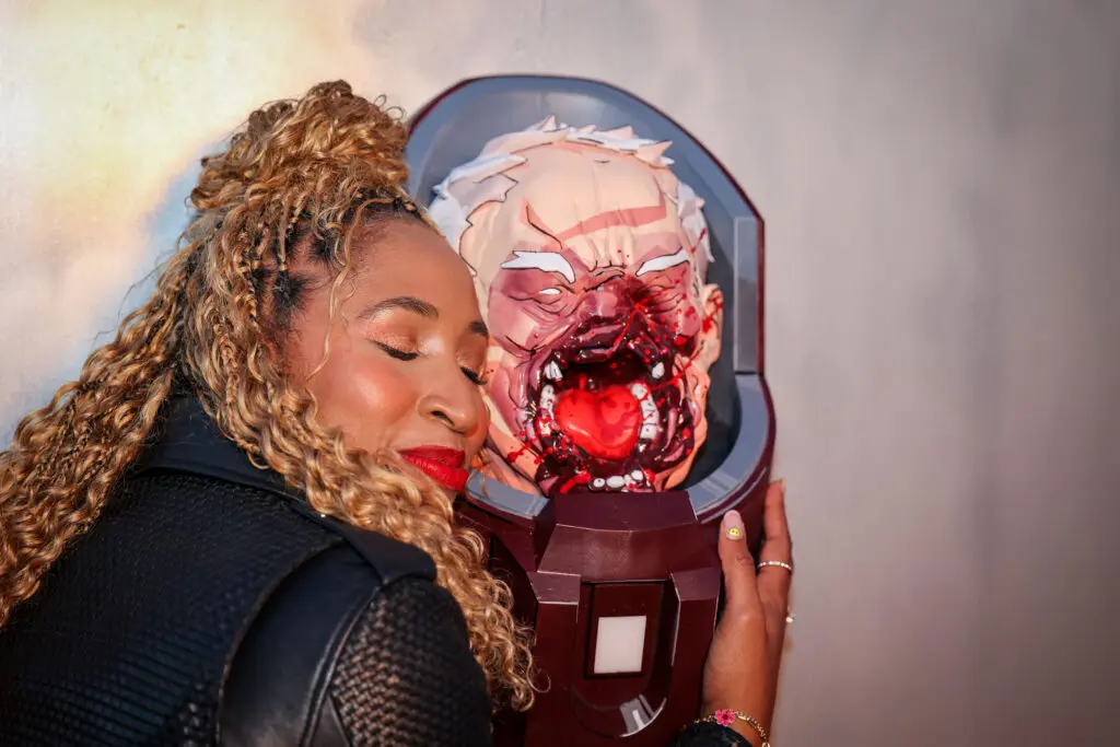A guest leans with closed eyes against a blood-splattered prosthetic Conquered Conquest head mounted on a bolted panel, glossy blood highlights and comic-style linework visible under warm event lighting.