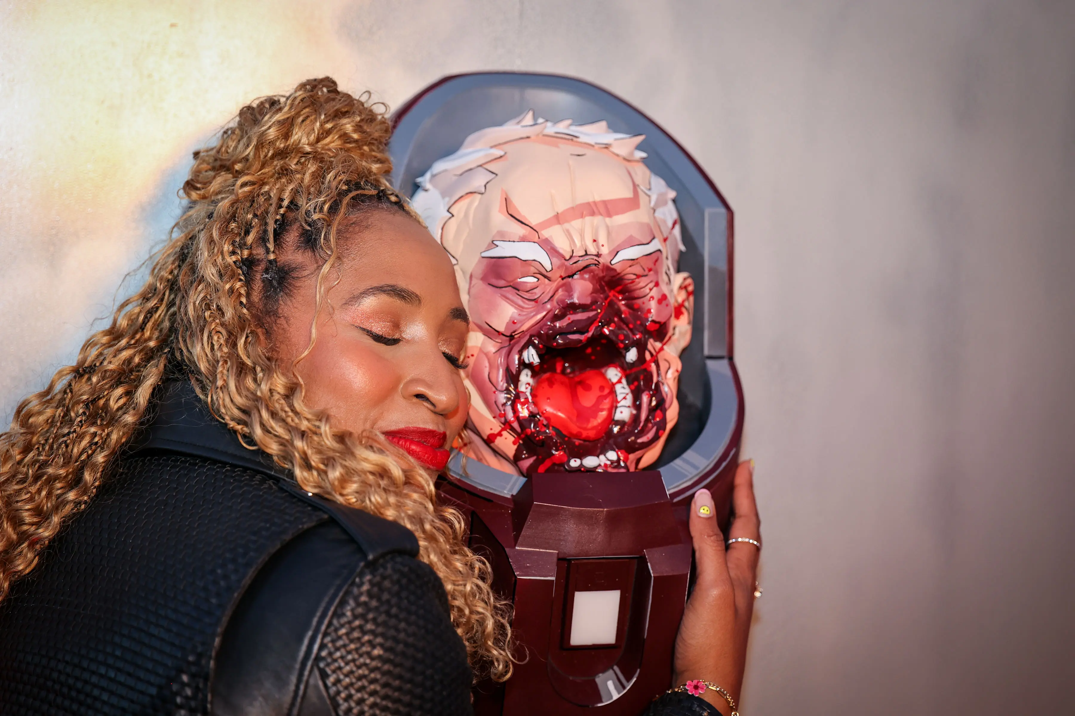 A guest leans with closed eyes against a blood-splattered prosthetic Conquered Conquest head mounted on a bolted panel, glossy blood highlights and comic-style linework visible under warm event lighting.