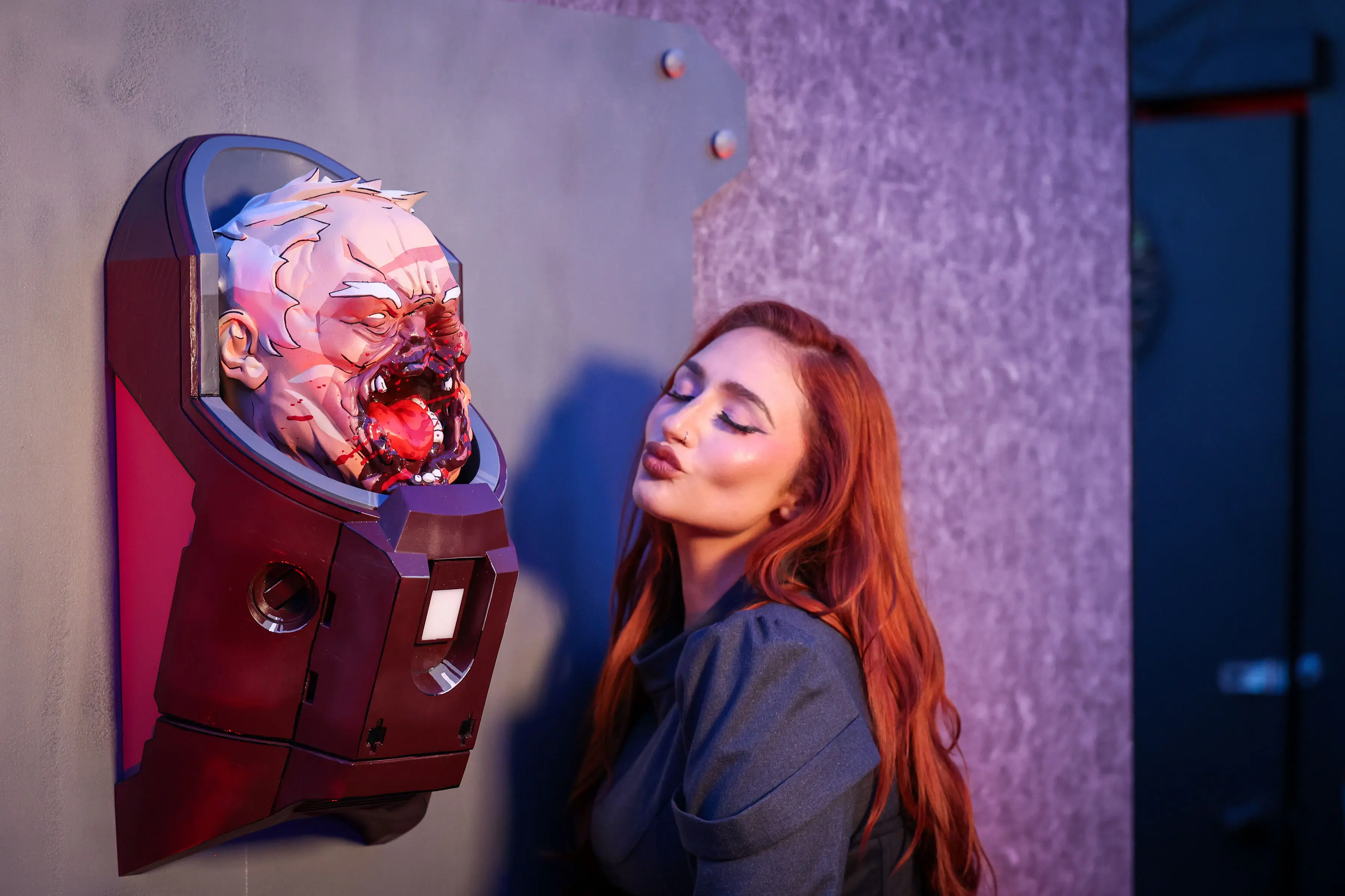 Blood-splattered, 3D-printed prosthetic head mounted on a bolted metal plate at left, with a red-haired creator puckering beside it under purple-blue event lighting that highlights glossy blood and ink linework.