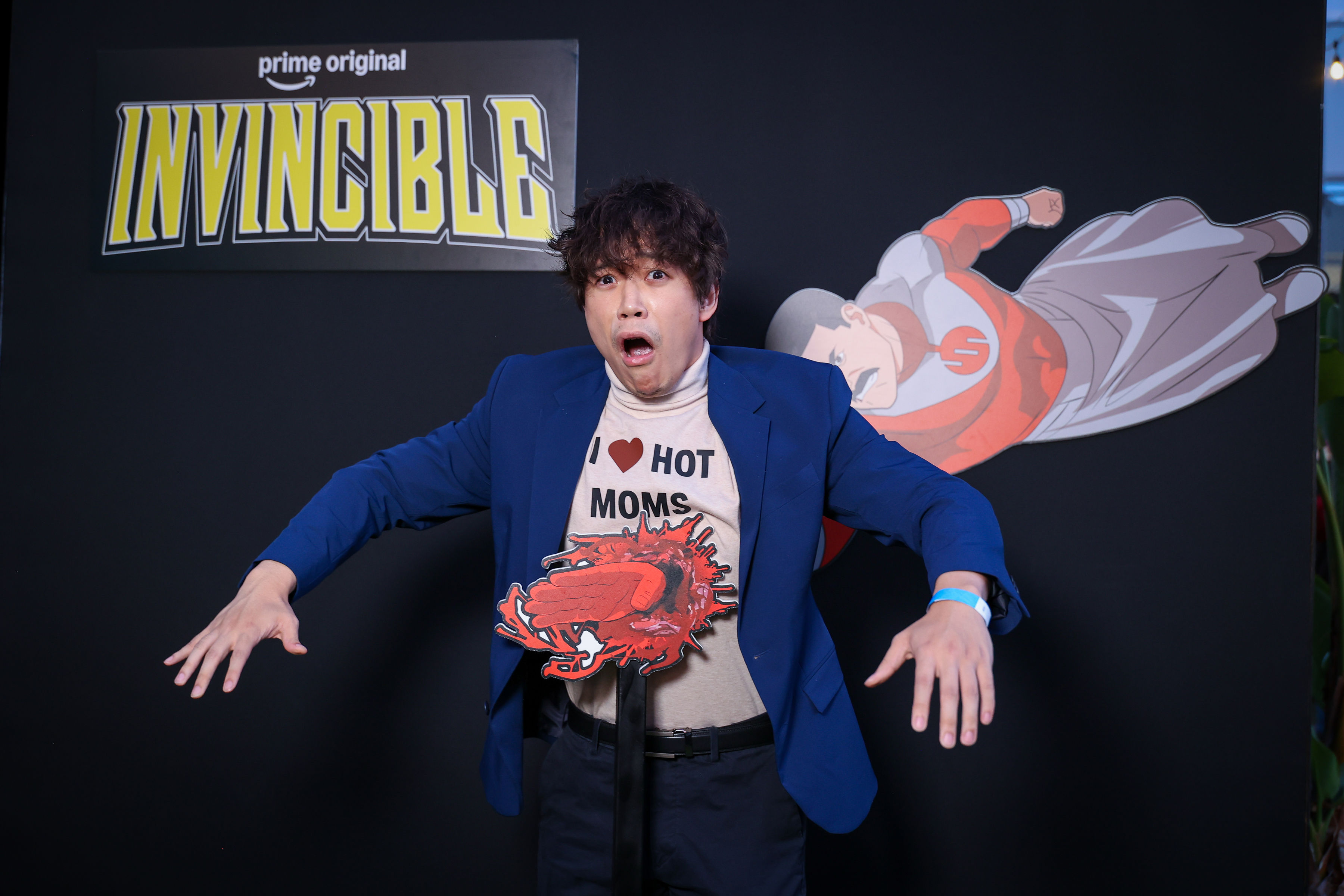 Person in a blue blazer makes a shocked face while posing against a black wall with an Invincible logo, a flying superhero cutout, and a red blood-splash prop at their abdomen.