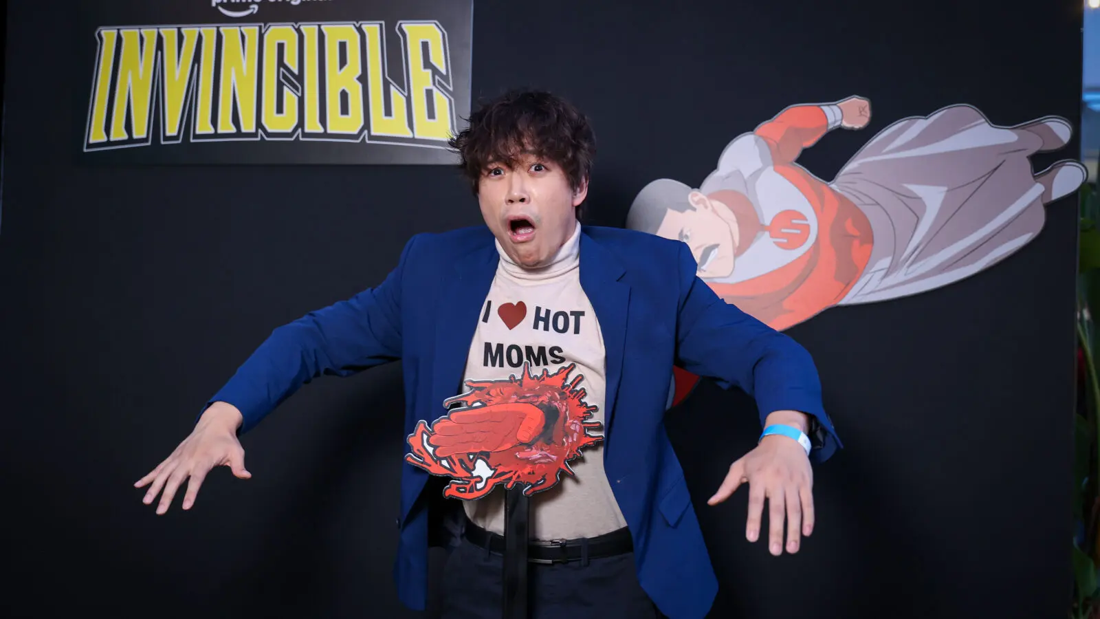 Person in a blue blazer and 'I ♥ HOT MOMS' shirt reacts with exaggerated shock in front of an 'Invincible' logo, posed with a blood-splatter prop and a flying superhero cutout on a black backdrop.