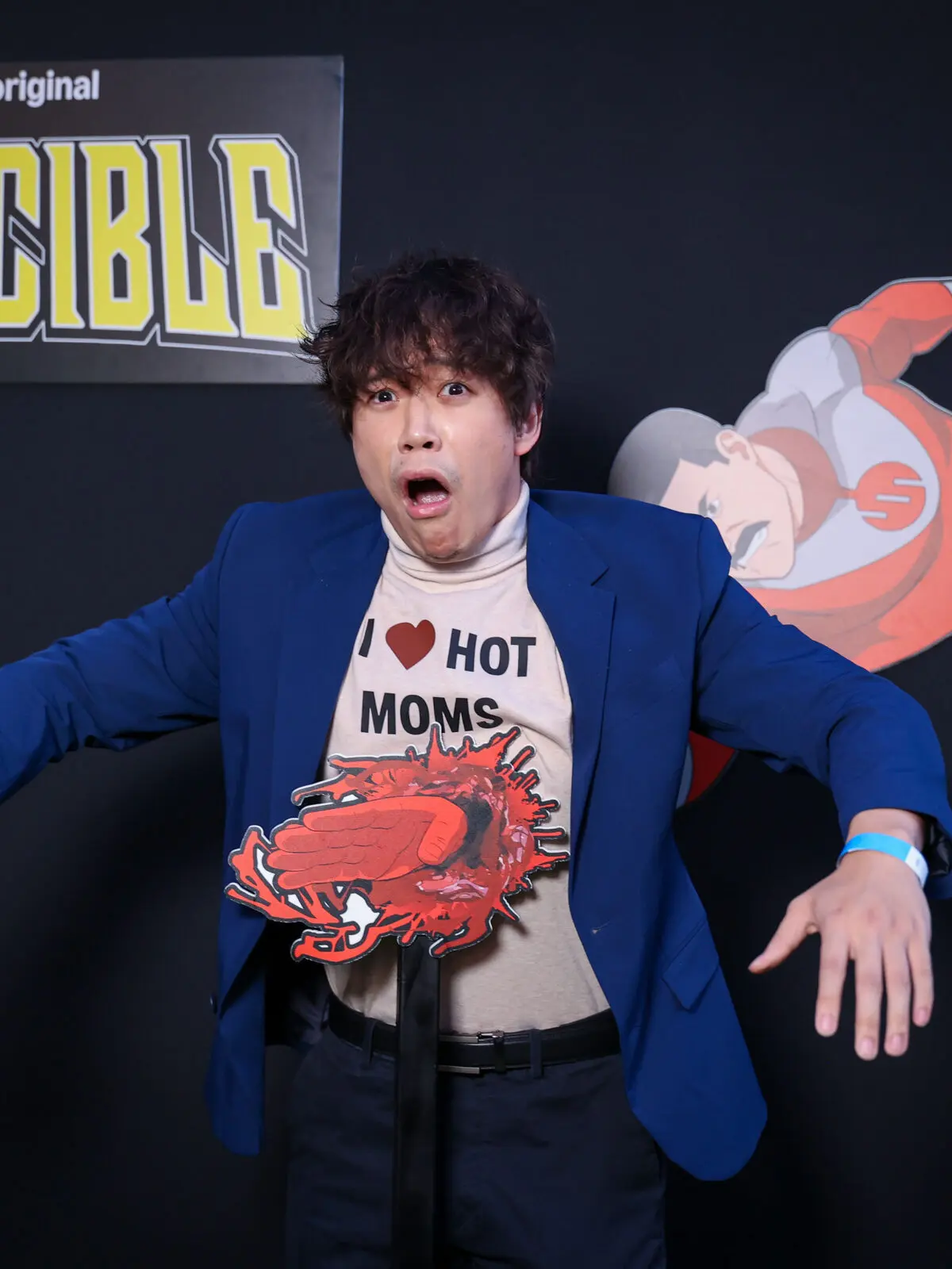 Person in a blue blazer and 'I ♥ HOT MOMS' shirt reacts with exaggerated shock in front of an 'Invincible' logo, posed with a blood-splatter prop and a flying superhero cutout on a black backdrop.
