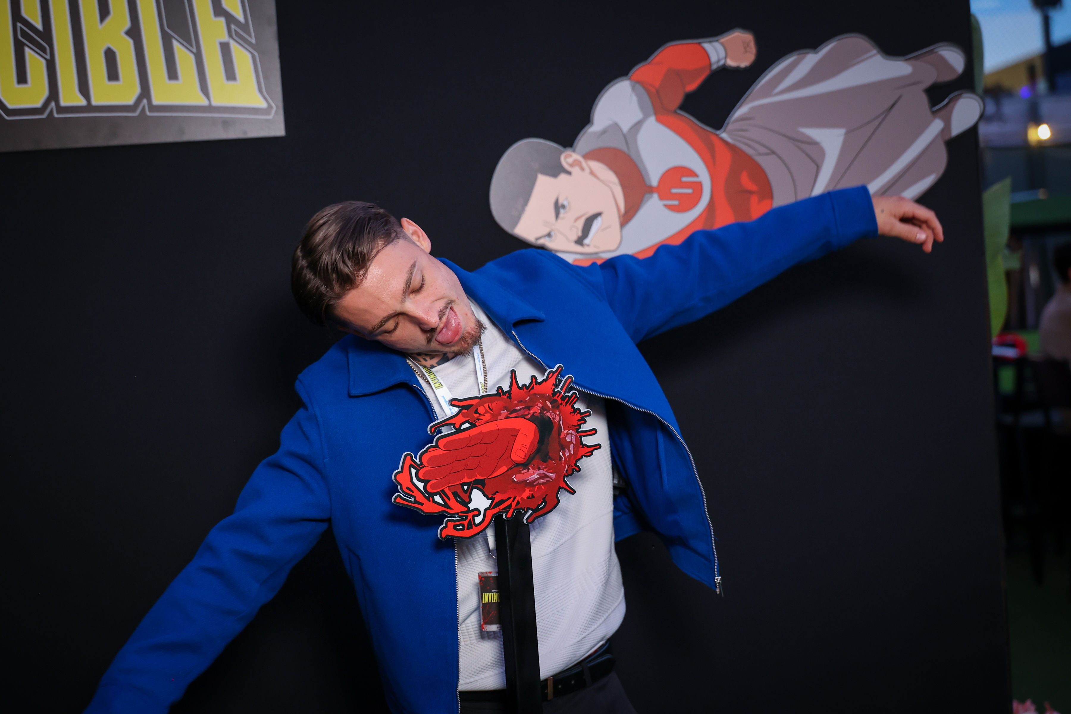 Man in a blue jacket pretends to be struck by a red blood-splash photo prop mounted on a stand in front of a black backdrop with a flying mustached superhero cutout and Invincible logo.