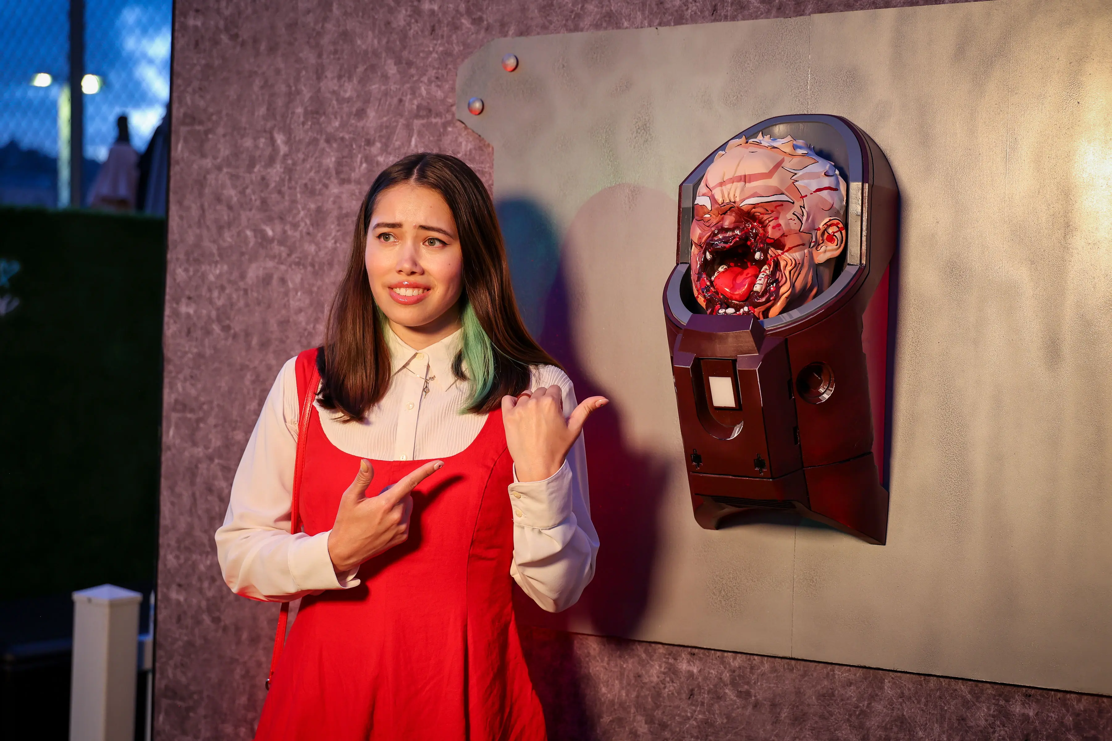 Young woman in a red dress gestures and points toward a bloodied, 3D-printed prosthetic head mounted on a bolted panel, lit by warm and purple event lighting for a creator activation.