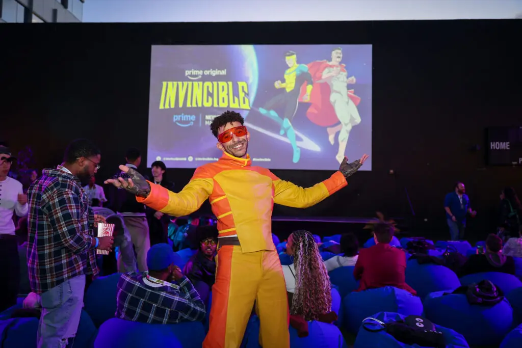 Khleo Thomas, dressed as Rex Splode in a yellow-and-orange costume, poses before a seated audience and projected Invincible artwork at the creator screening.