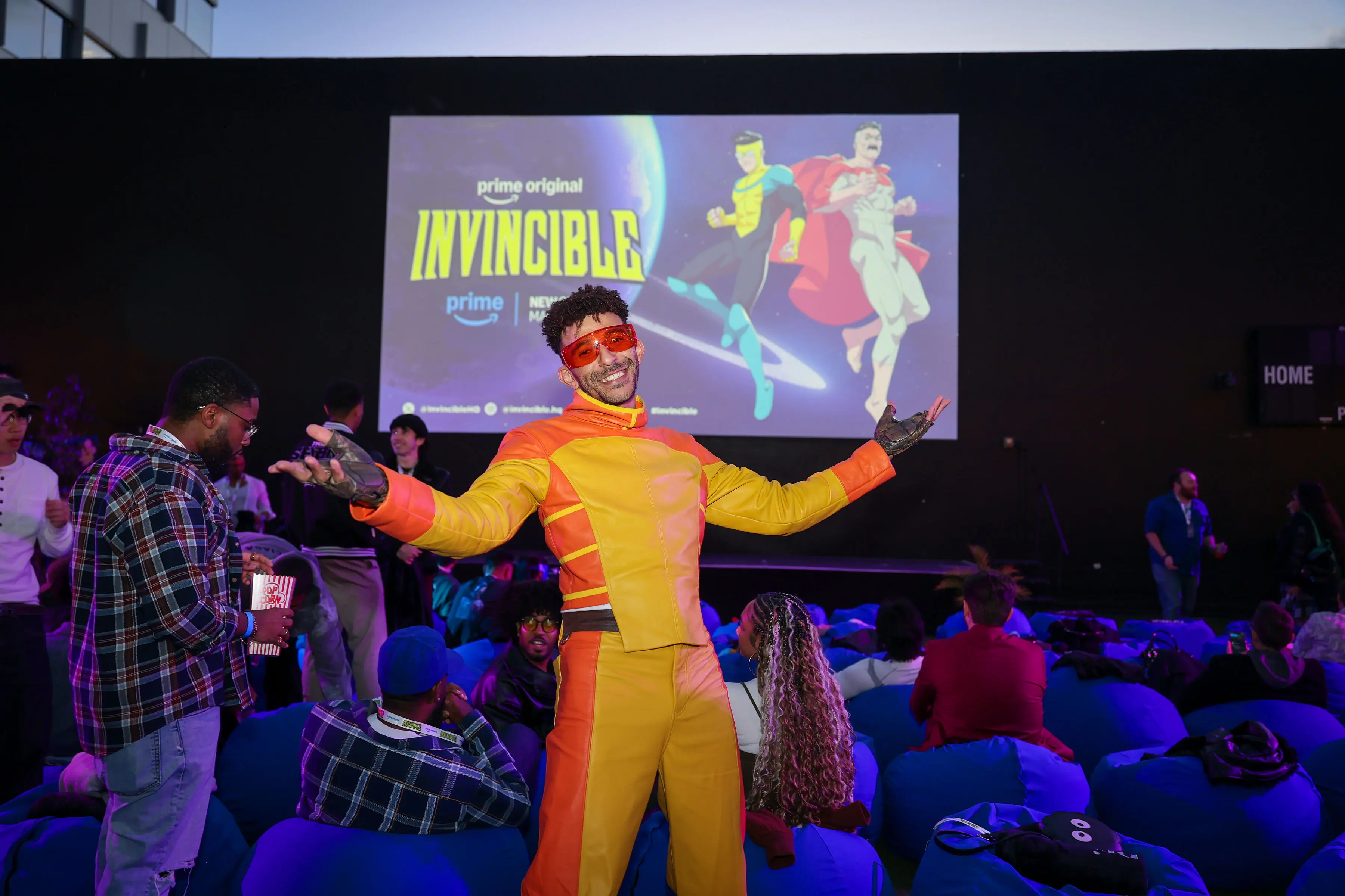 Costumed creator in a yellow-and-orange suit and orange visor gestures to the camera in front of a large projected INVINCIBLE wordmark and hero art, with beanbag seating and an audience behind him.