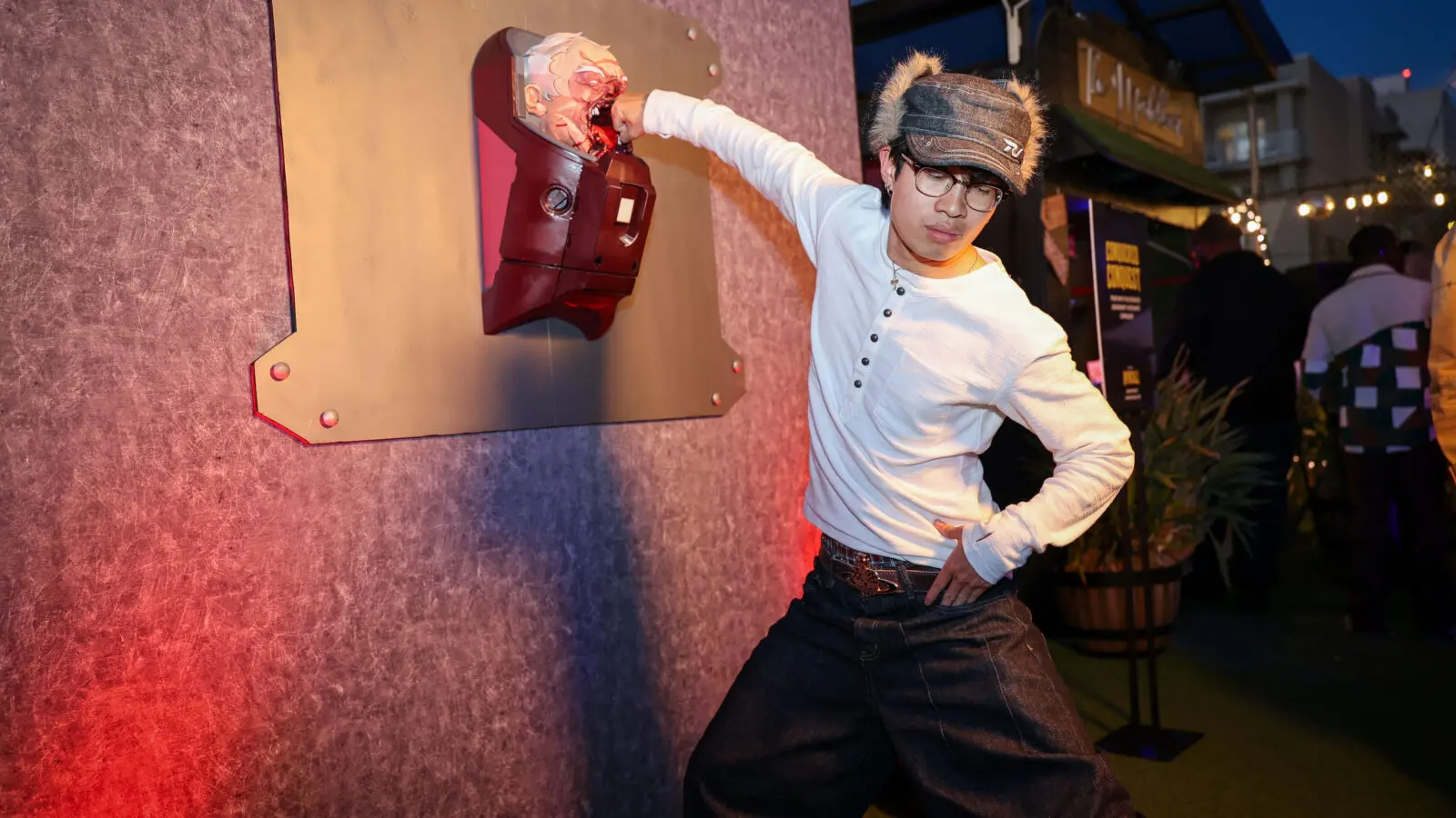 Guest poses punching a bloodied, mounted superhero head prop fixed to a textured wall at an evening Invincible Season 4 event, lit by warm red and cool blue party lights.