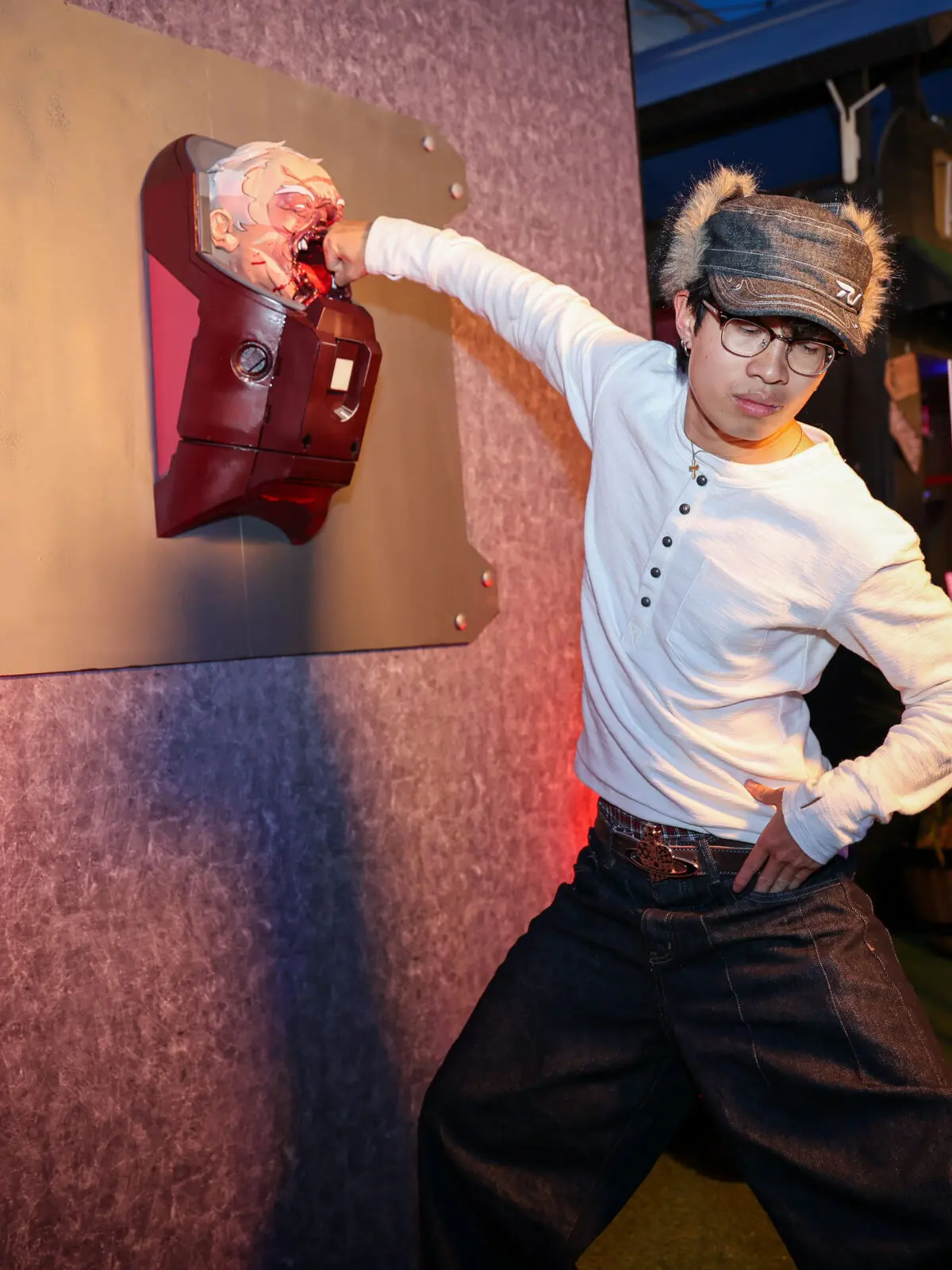 Guest poses punching a bloodied, mounted superhero head prop fixed to a textured wall at an evening Invincible Season 4 event, lit by warm red and cool blue party lights.