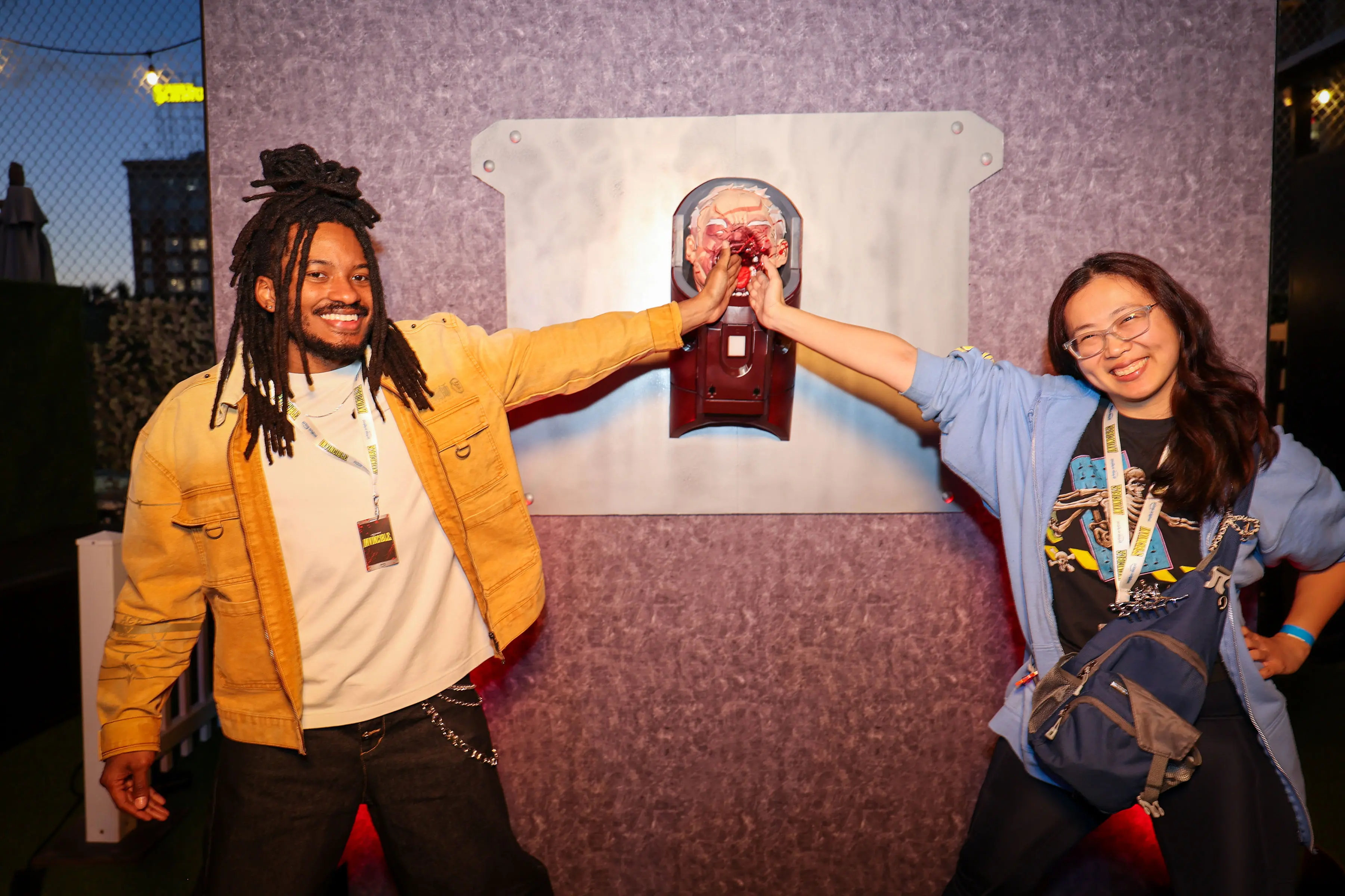 Two smiling event attendees press their palms against a blood-splattered prosthetic head mounted on a purple textured panel, wearing Invincible lanyards and casual jackets while posing for an interactive photo moment.