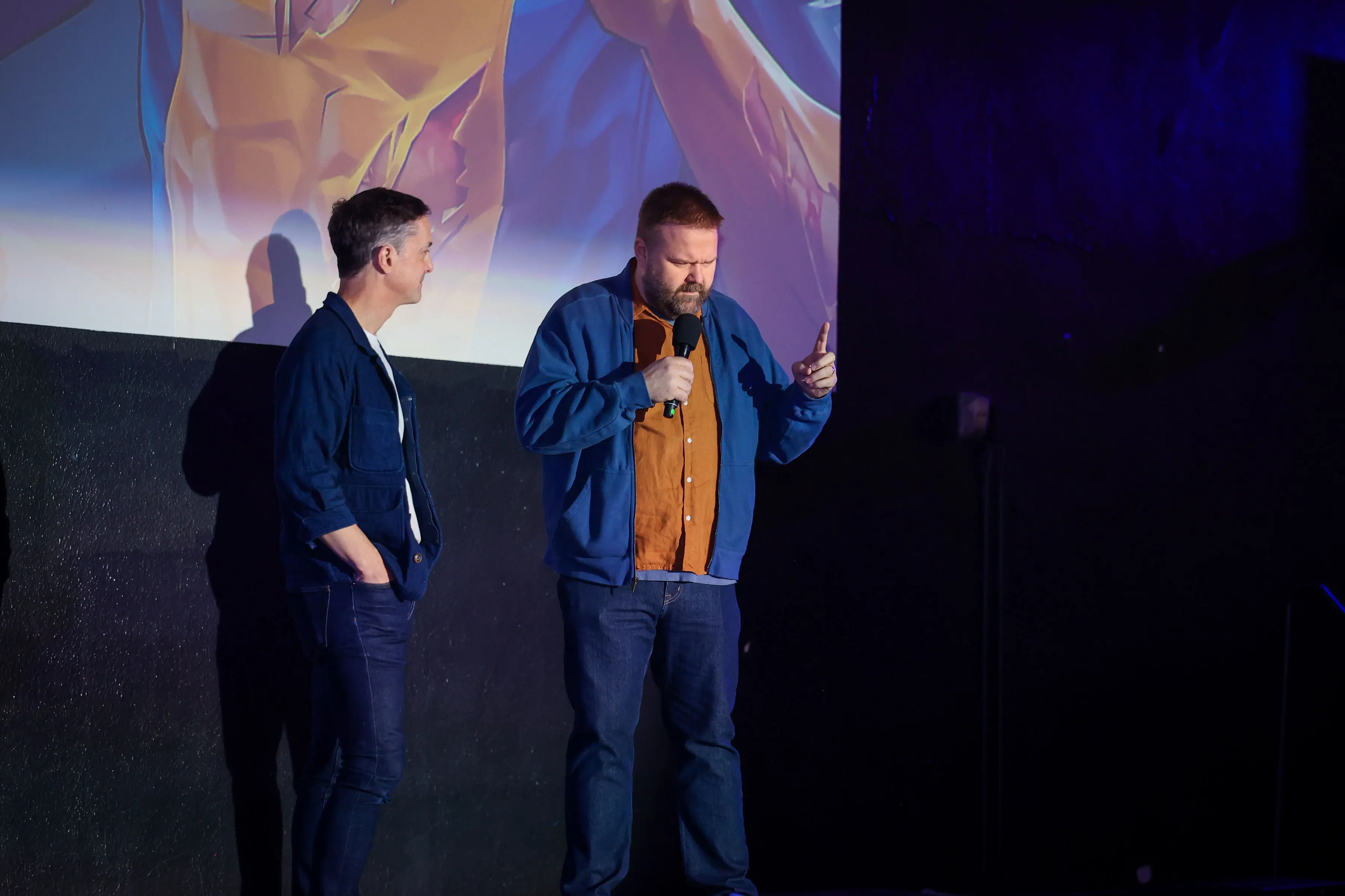 Two men on a dim stage, one holding a microphone and mid-gesture beneath a large projected yellow-and-blue Invincible illustration, blue and purple stage lighting and deep shadows.