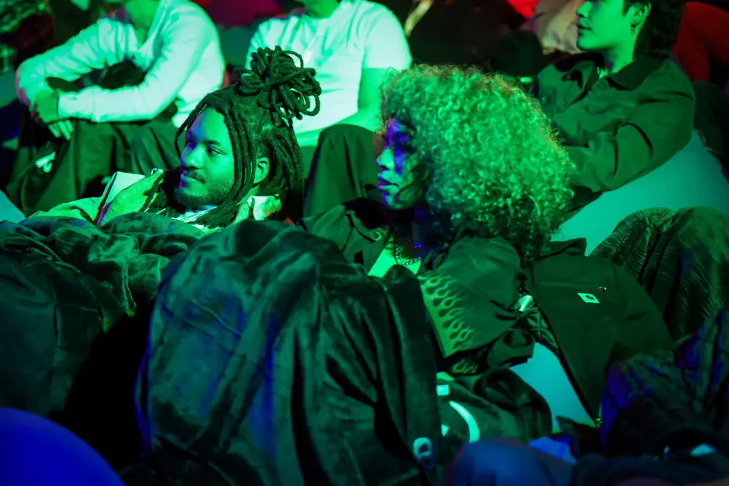 Two audience members recline in blanket-covered beanbags, bathed in saturated green and purple screening light, watching the Invincible Season 4 creator screening while other attendees sit behind them.