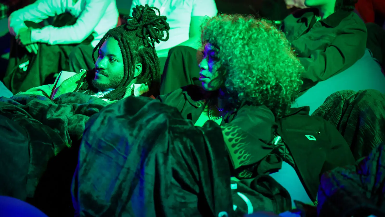 Two people reclining on beanbag seating under saturated green stage light, wrapped in blankets with other attendees visible behind them at a dim, immersive screening event.